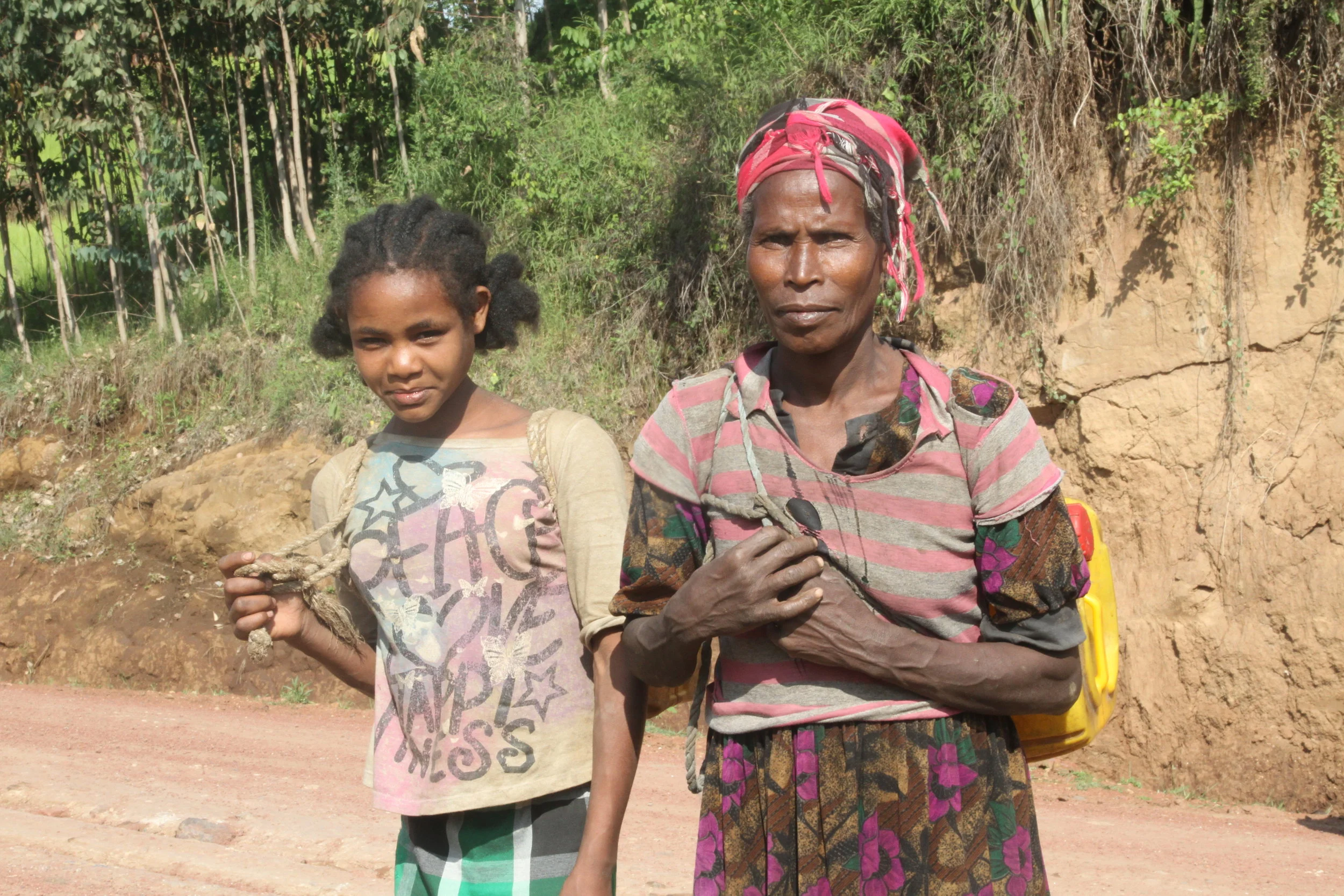 A mom and daughter carry 40 lb jerry cans on their backs with improvised rope harness.