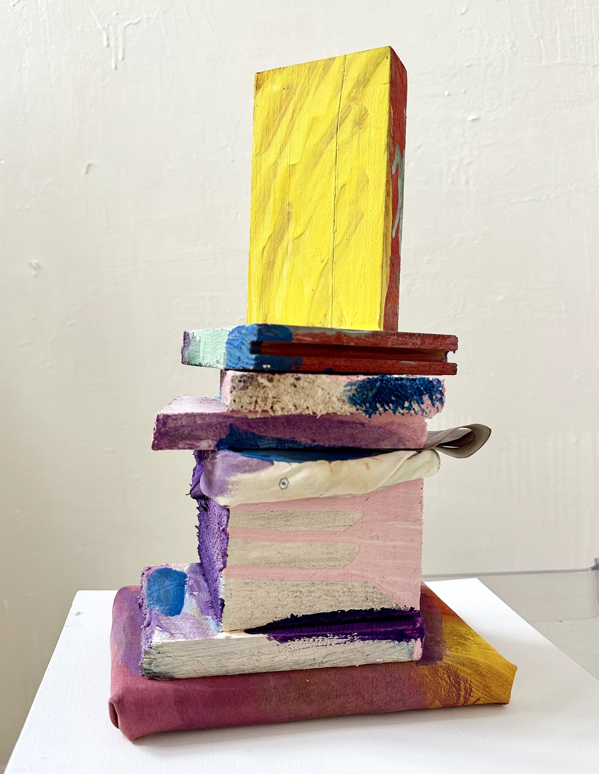 Abstract sculpture made of stacked, painted, and wrapped objects, including books and blocks, with bright colors like yellow, pink, purple, blue, and black, against a plain white wall.