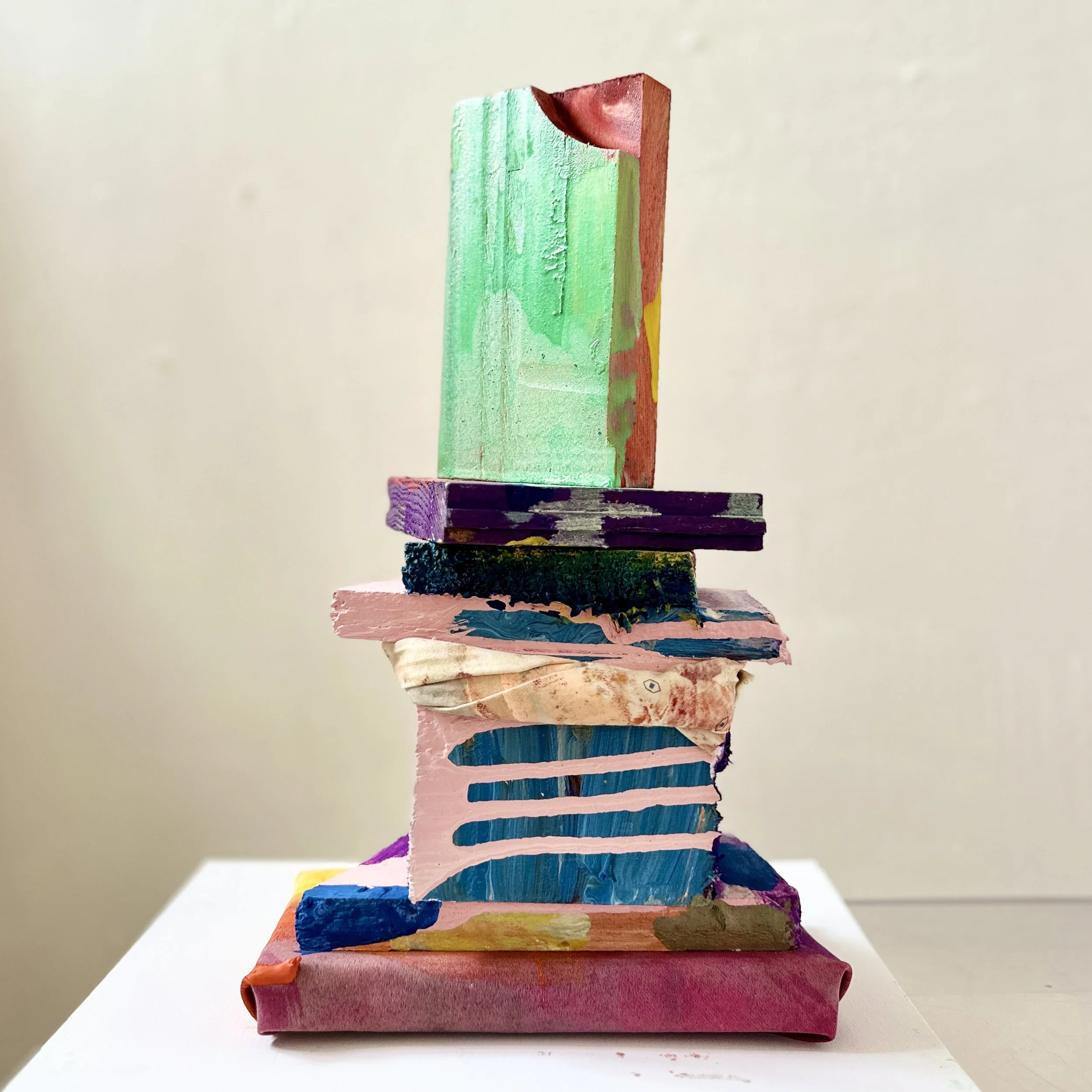 Colorful abstract sculpture made of stacked painted wooden blocks, with various patterns and textures, on a white surface against a plain wall.