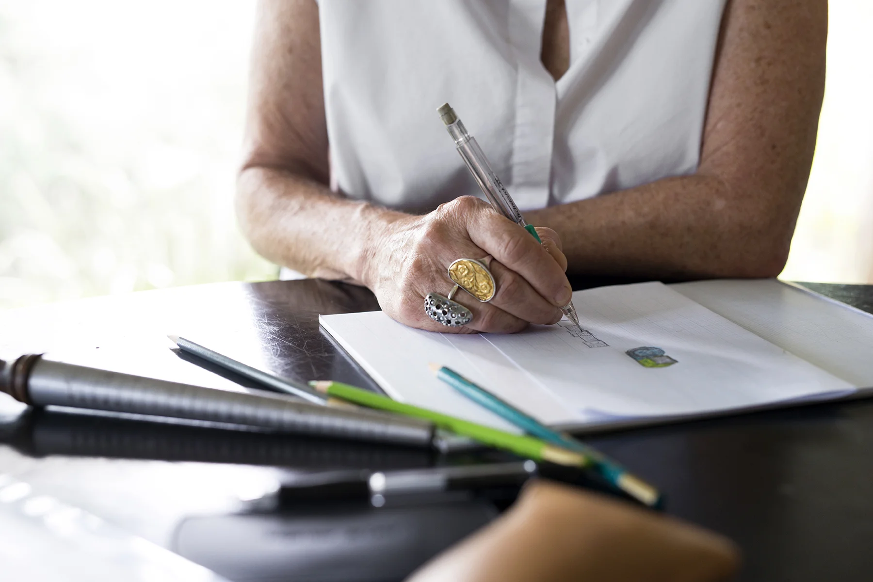 Barb sketching some ring designs. Natural light fills the space from the wall of windows behind her.