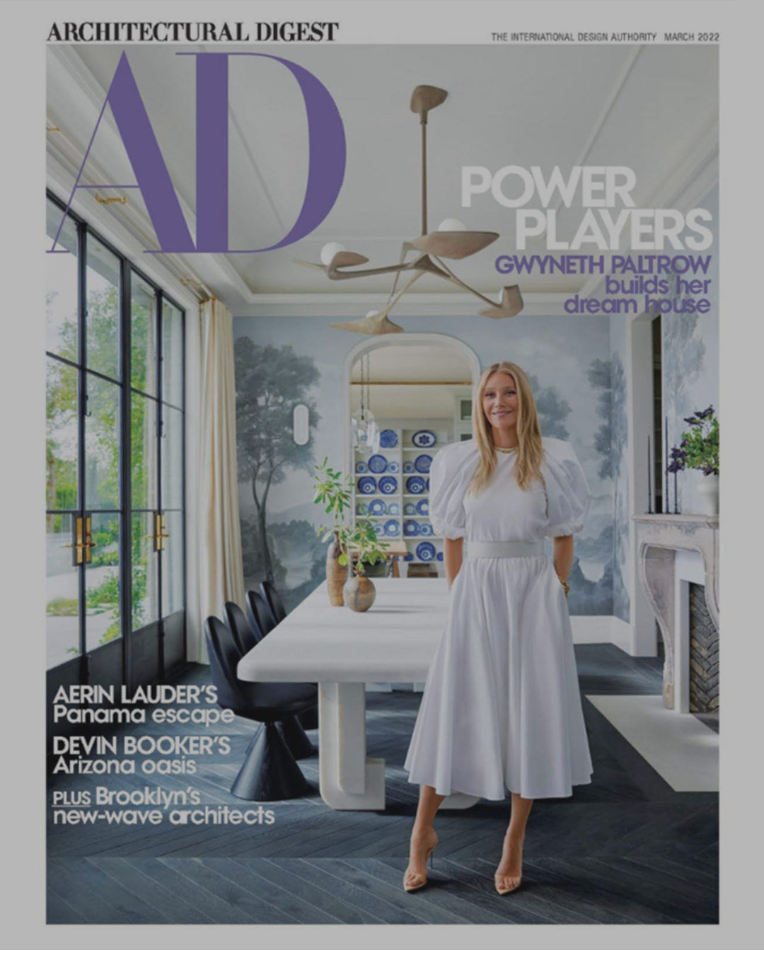 Cover of Architectural Digest magazine featuring a woman in a white dress standing in a modern dining room with a large table, decorative chandelier, and wall art. Devon Grace Interiors, DGI design build, interior design and design build in chicago I