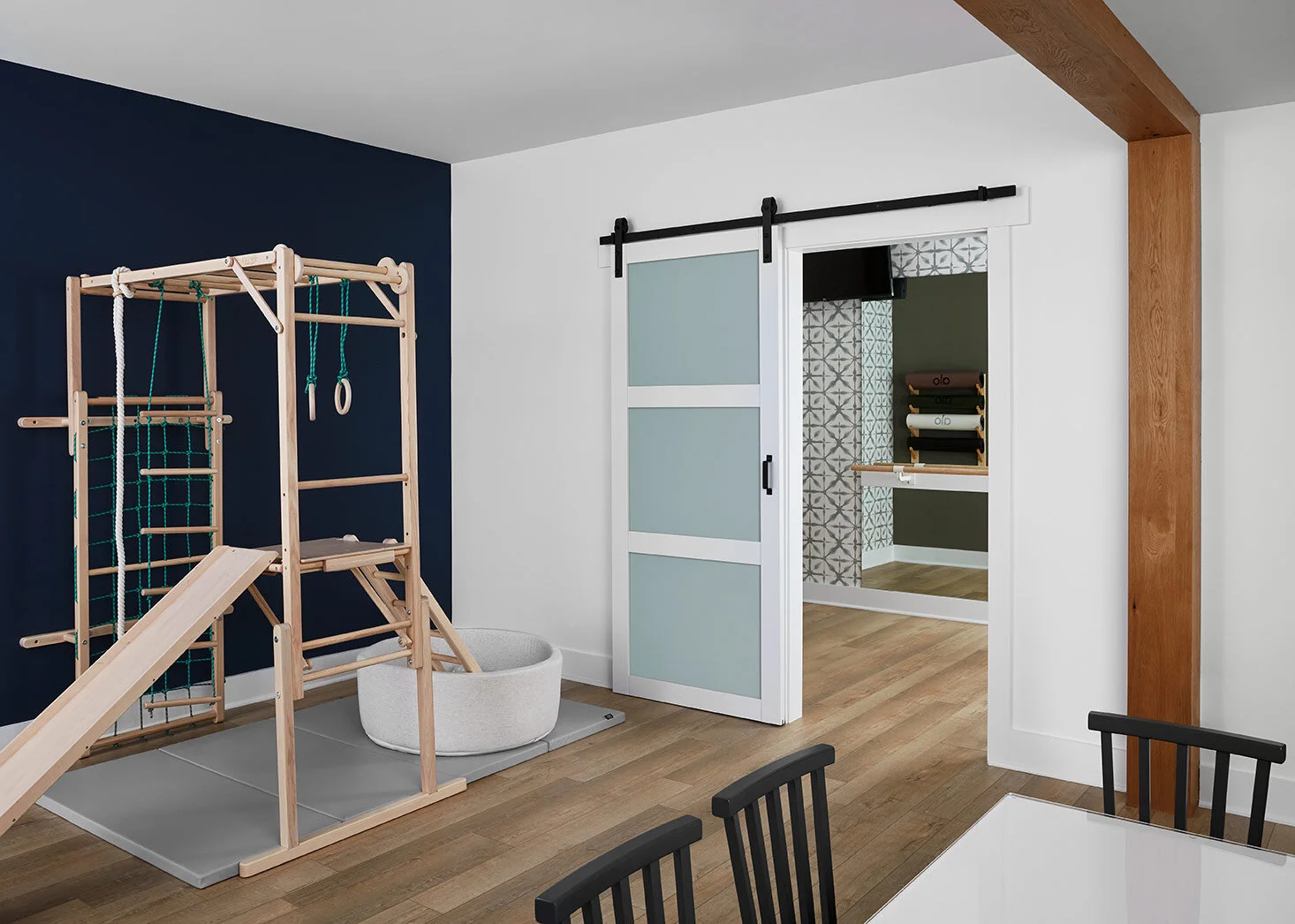 Behind the family room area, the kids get their own space to enjoy! A playground anchors the space, along with an arts and crafts table. Behind the kids area, a barn door separates a small home gym which was celebrated with a fun wallpaper to add so…