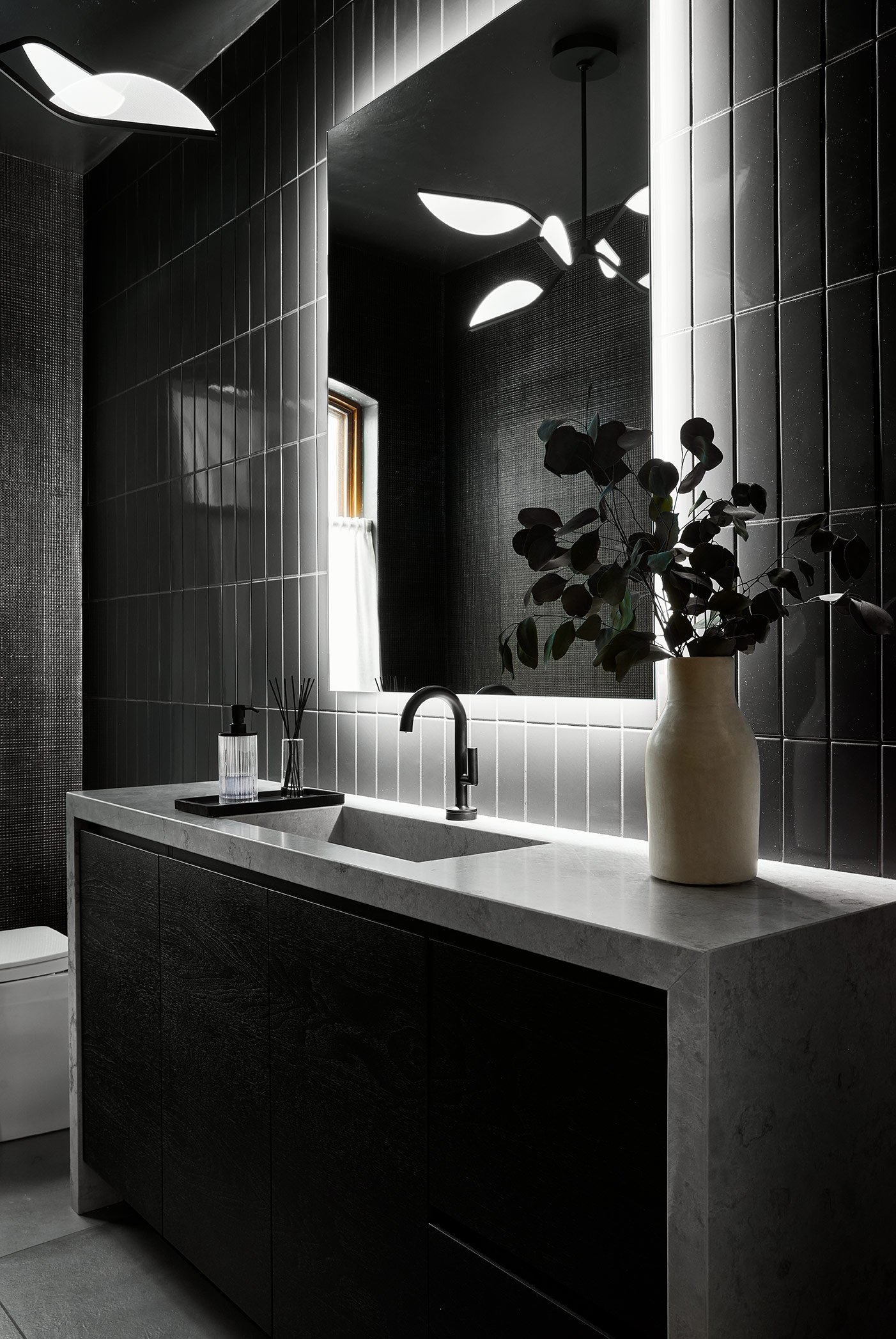 Black bathroom cabinet with gray marble countertop and exterior. Light gray floor tiles and backlit mirror.