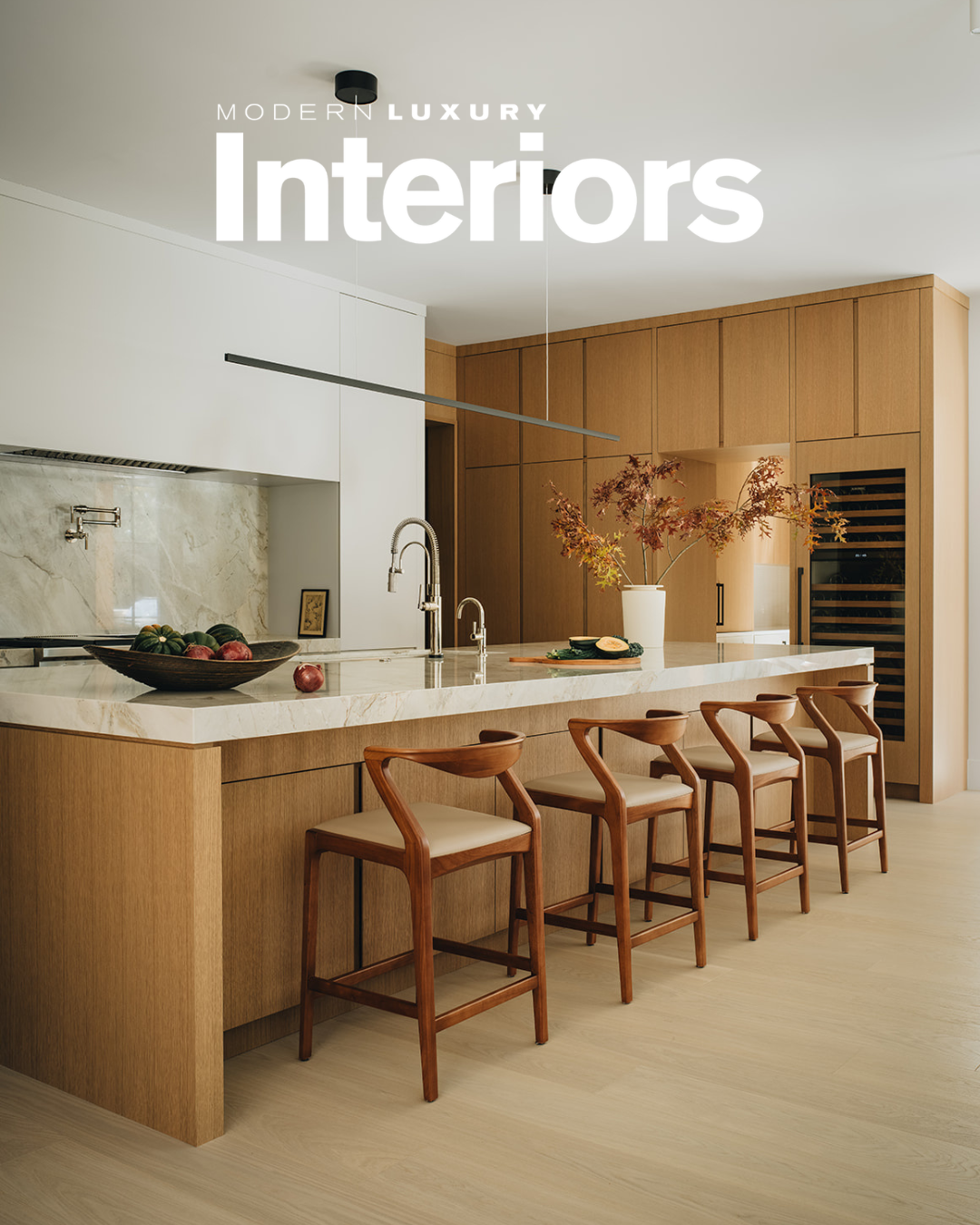Modern kitchen with wooden cabinets, a marble countertop, and four wooden bar stools with beige cushions. There is a bowl of fruit, a vase with fall leaves, and a sink with a high-arc faucet.