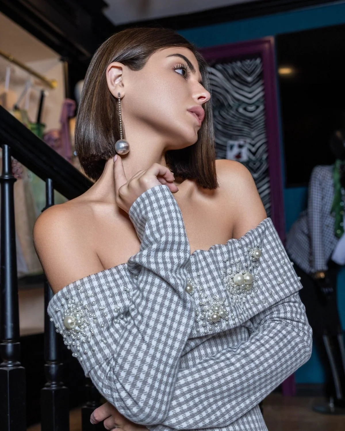 A woman with shoulder-length dark hair, wearing a checkered off-shoulder dress with pearl embellishments, is touching her neck and looking to the side. She has a long, dangling pearl earring.