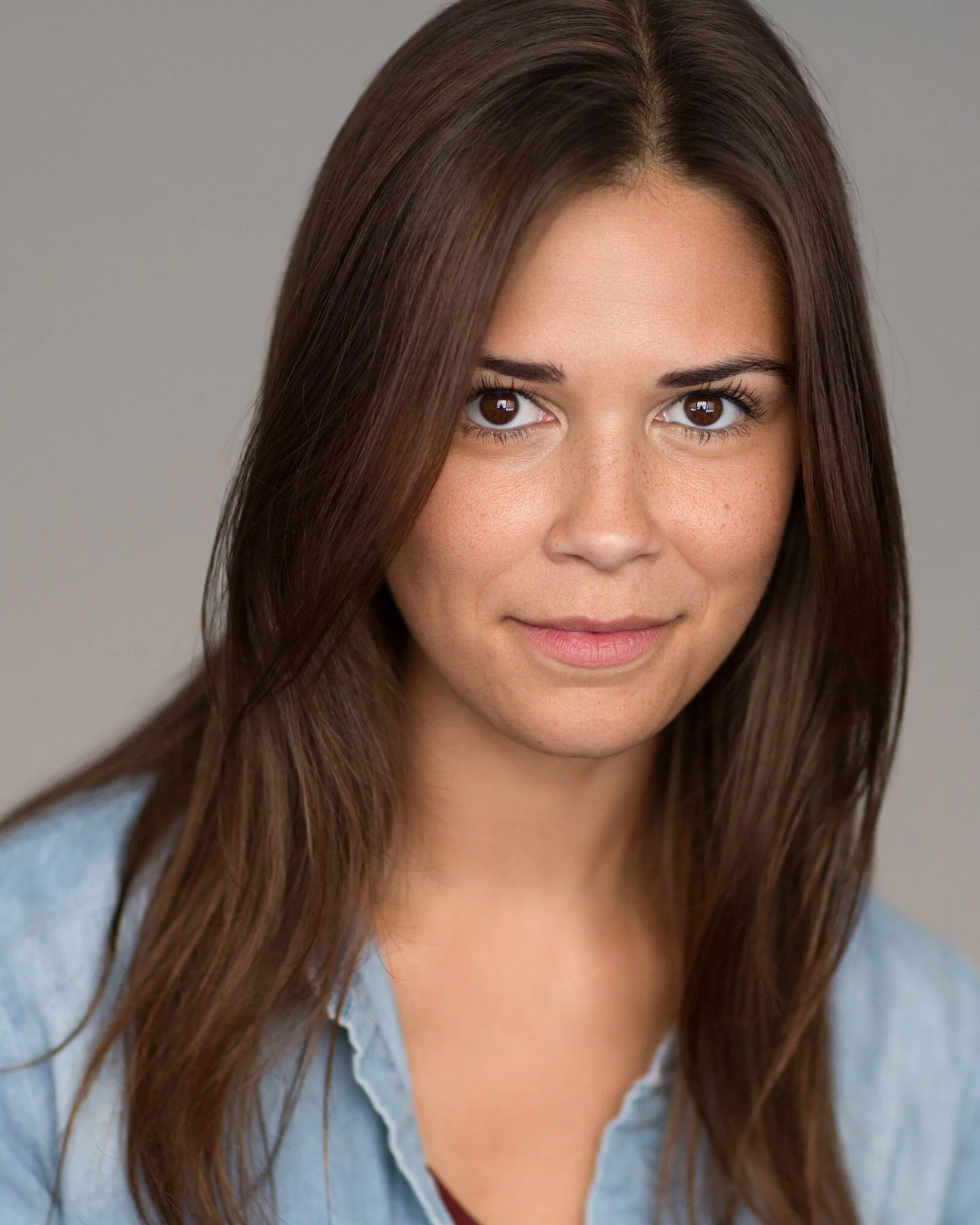 A woman with long brown hair and light skin, wearing a denim shirt, looking at the camera with a slight smile.