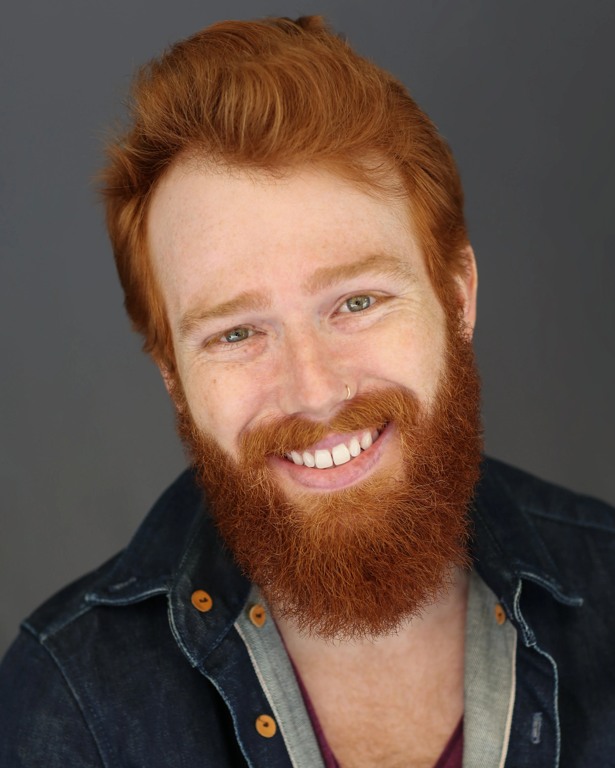 Smiling man with red hair and beard, wearing a denim jacket and gray shirt, against a gray background.