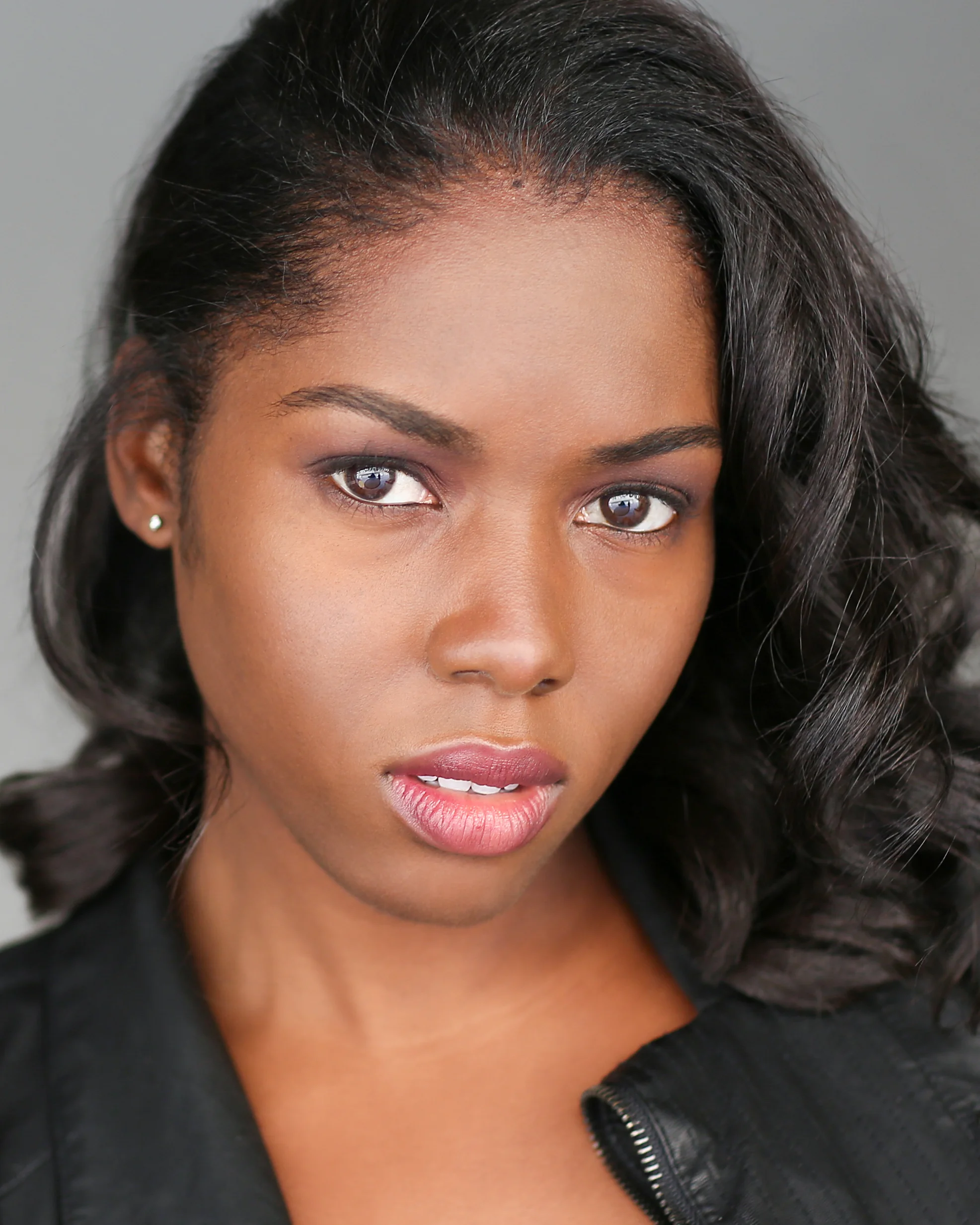 Close-up of a woman with dark wavy hair, light makeup, earrings, wearing a black leather jacket against a gray background.