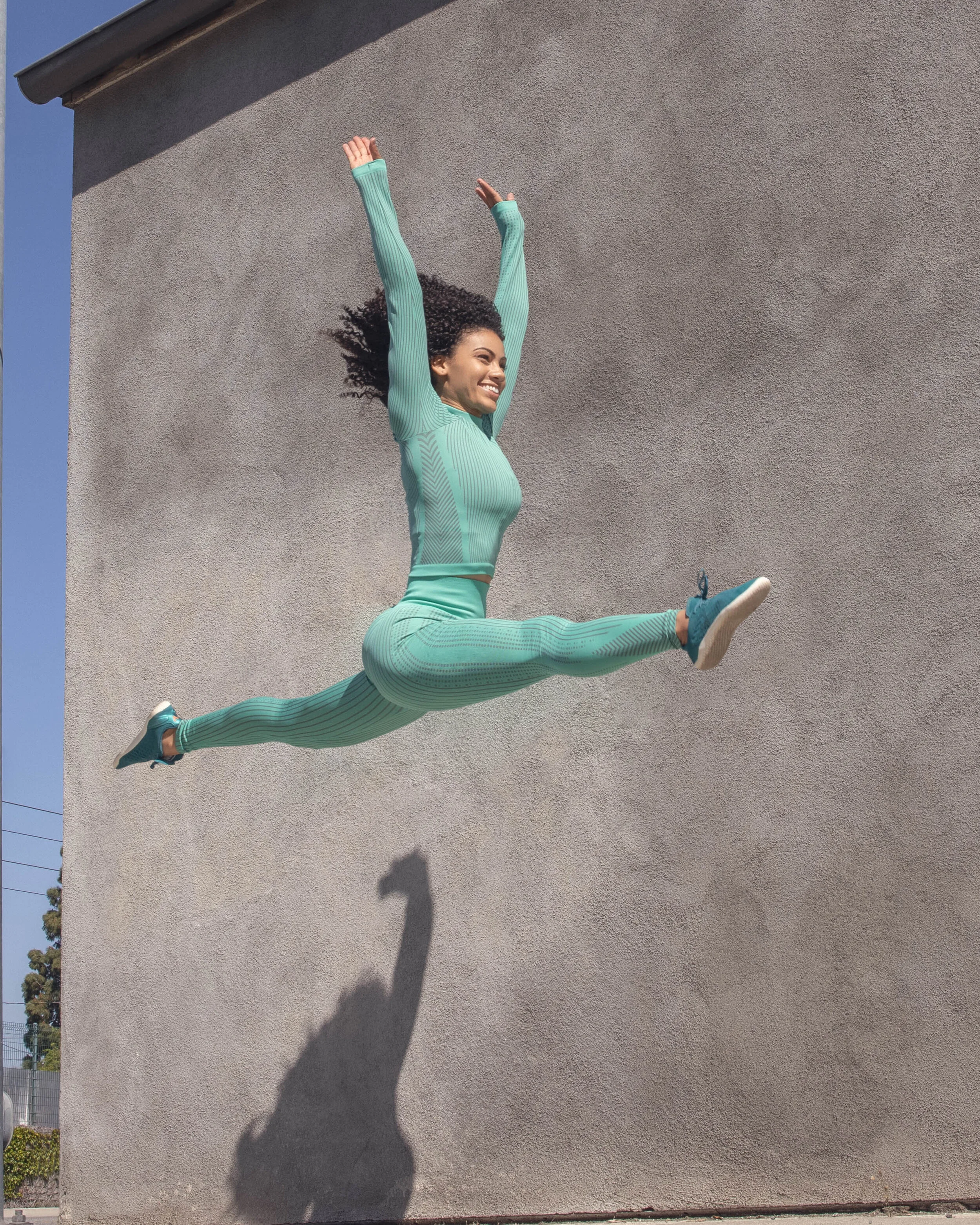 A woman jumping with arms raised and legs extended against a textured gray wall, casting a shadow on the wall.
