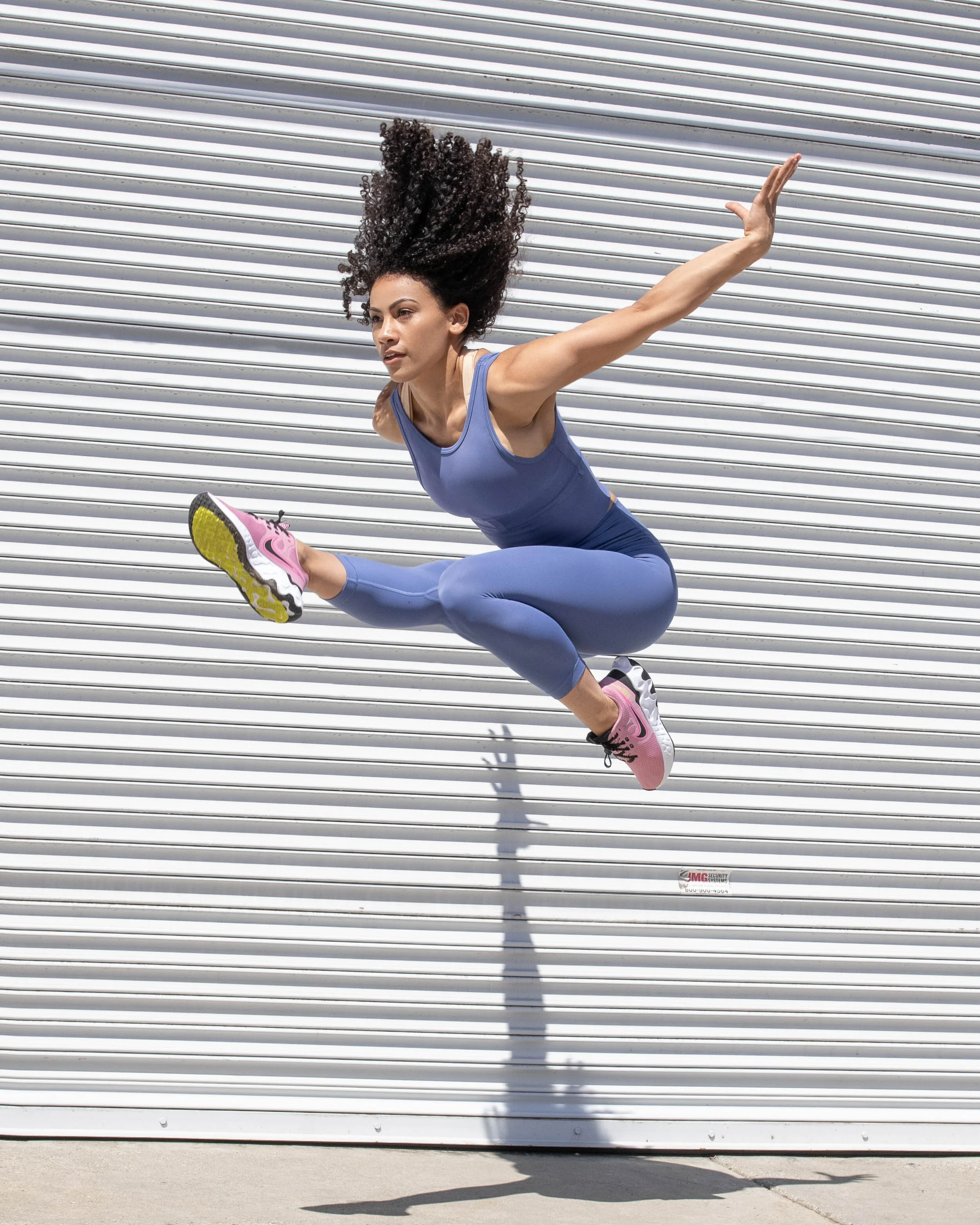 A woman in a blue athletic outfit performing a mid-air jump in front of a metal garage door.