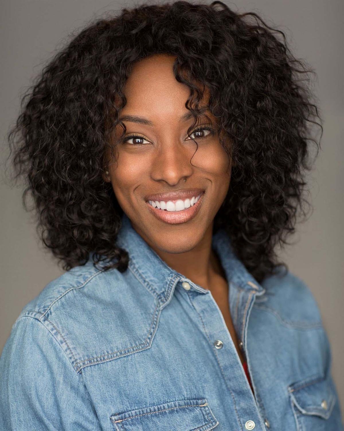 Portrait of a smiling woman with curly black hair, wearing a denim shirt against a plain background.