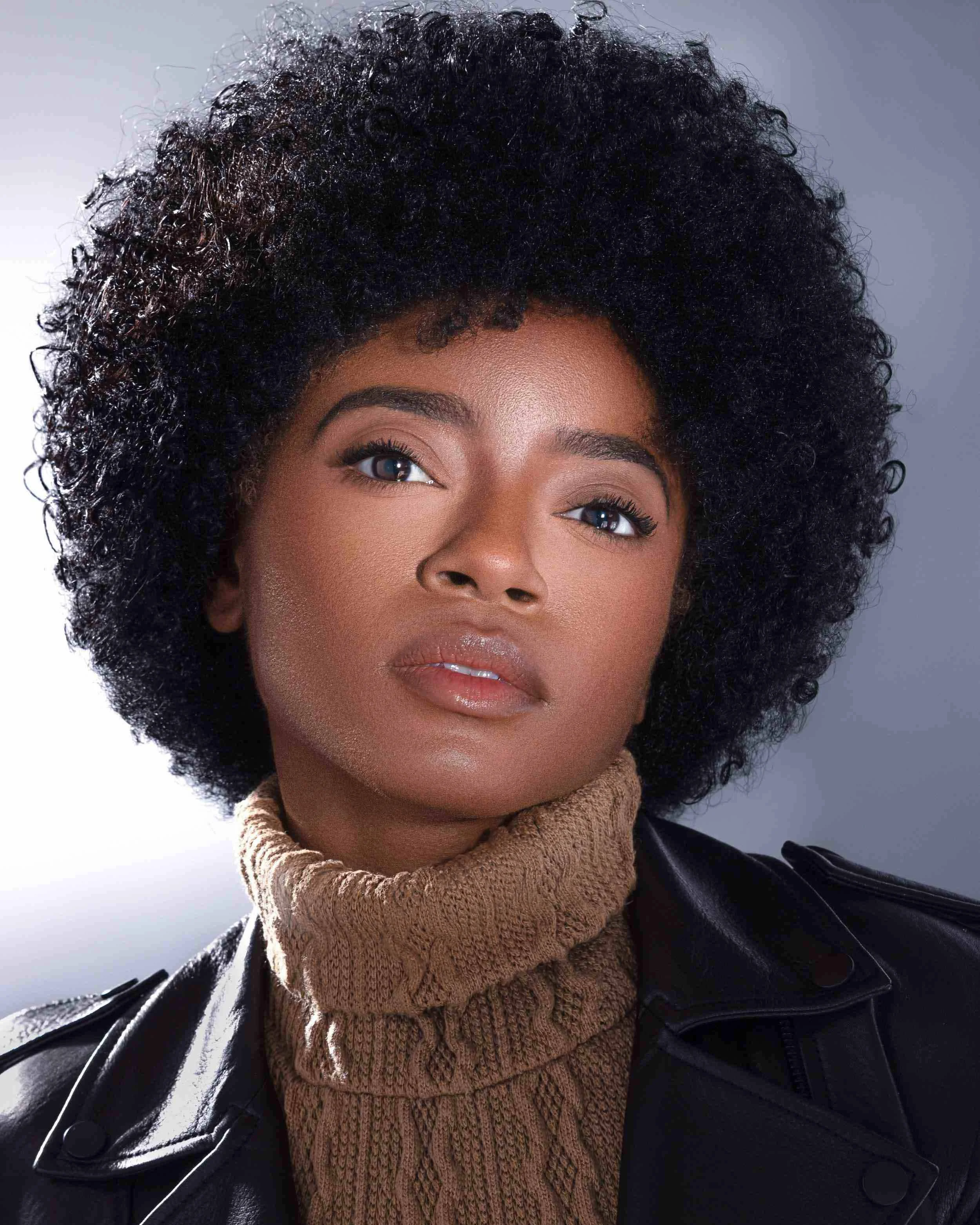 A woman with a voluminous natural afro hairstyle, wearing a tan turtleneck sweater and black leather jacket, staring confidently at the camera against a neutral background.
