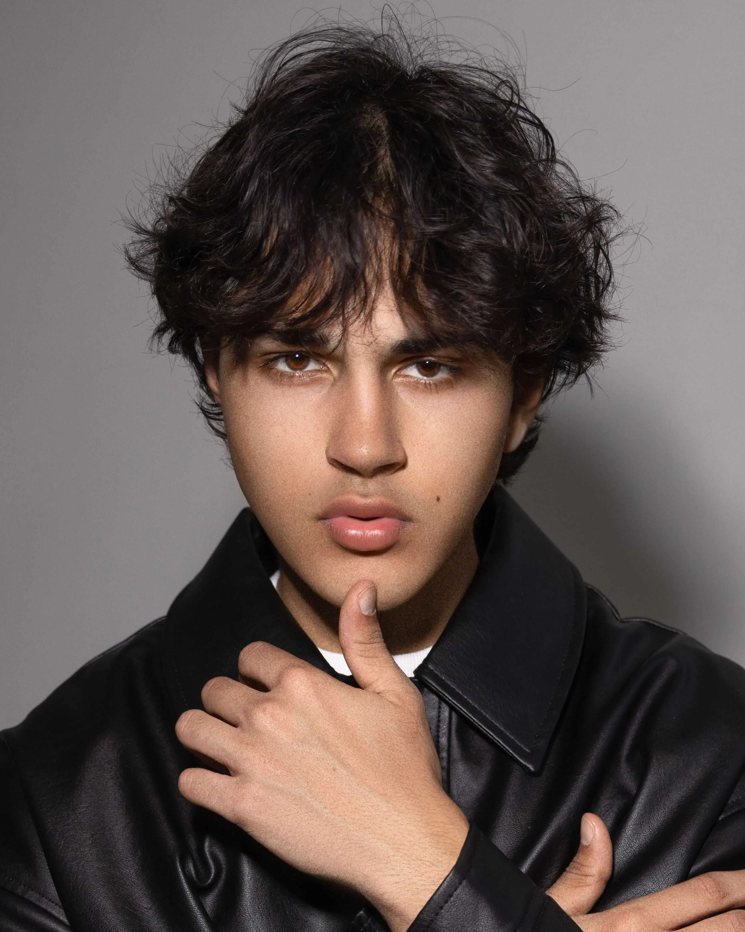 Young man with wavy dark hair wearing a black leather jacket posing against a plain background.