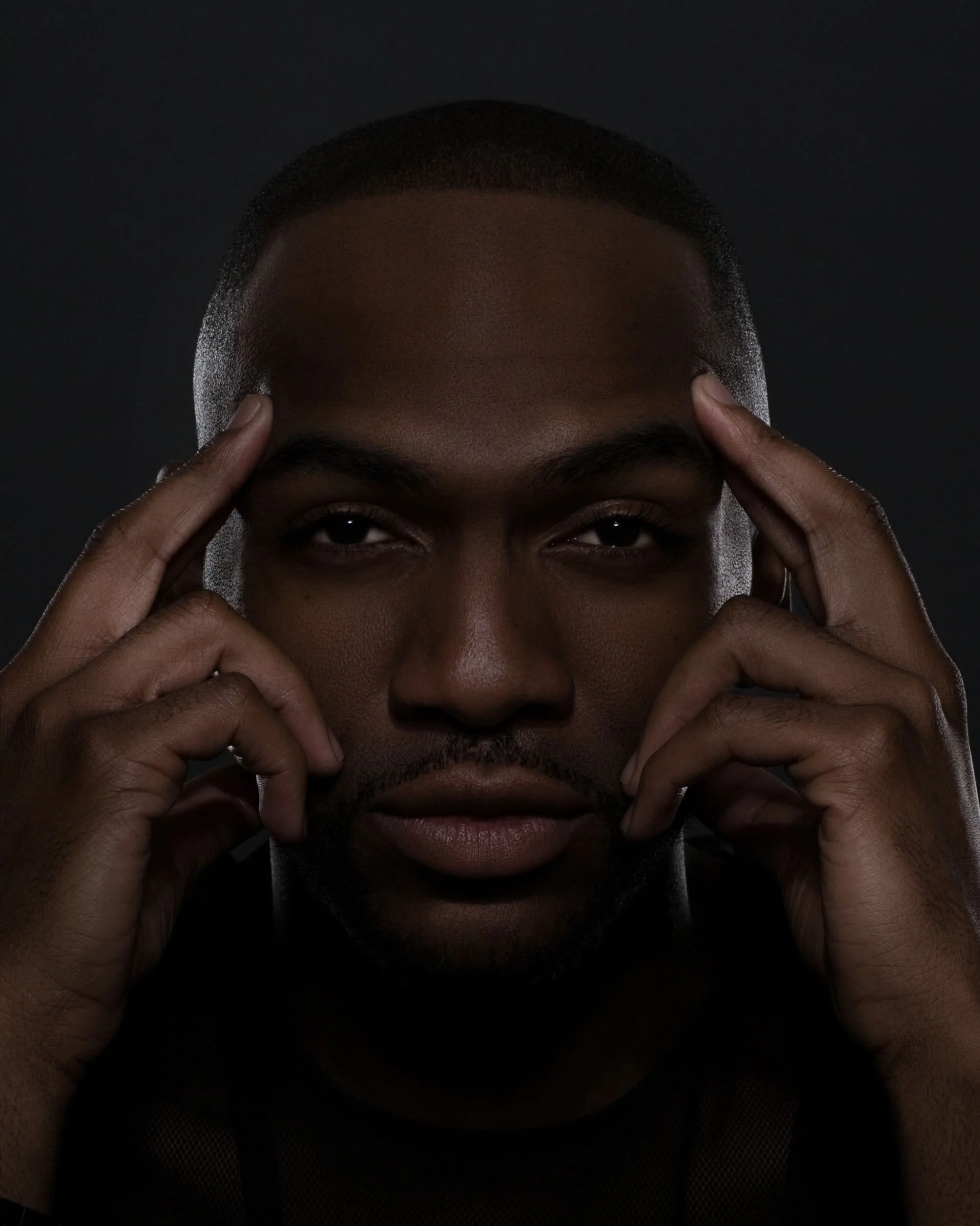 Close-up portrait of a man touching his temples with both hands against a dark background.