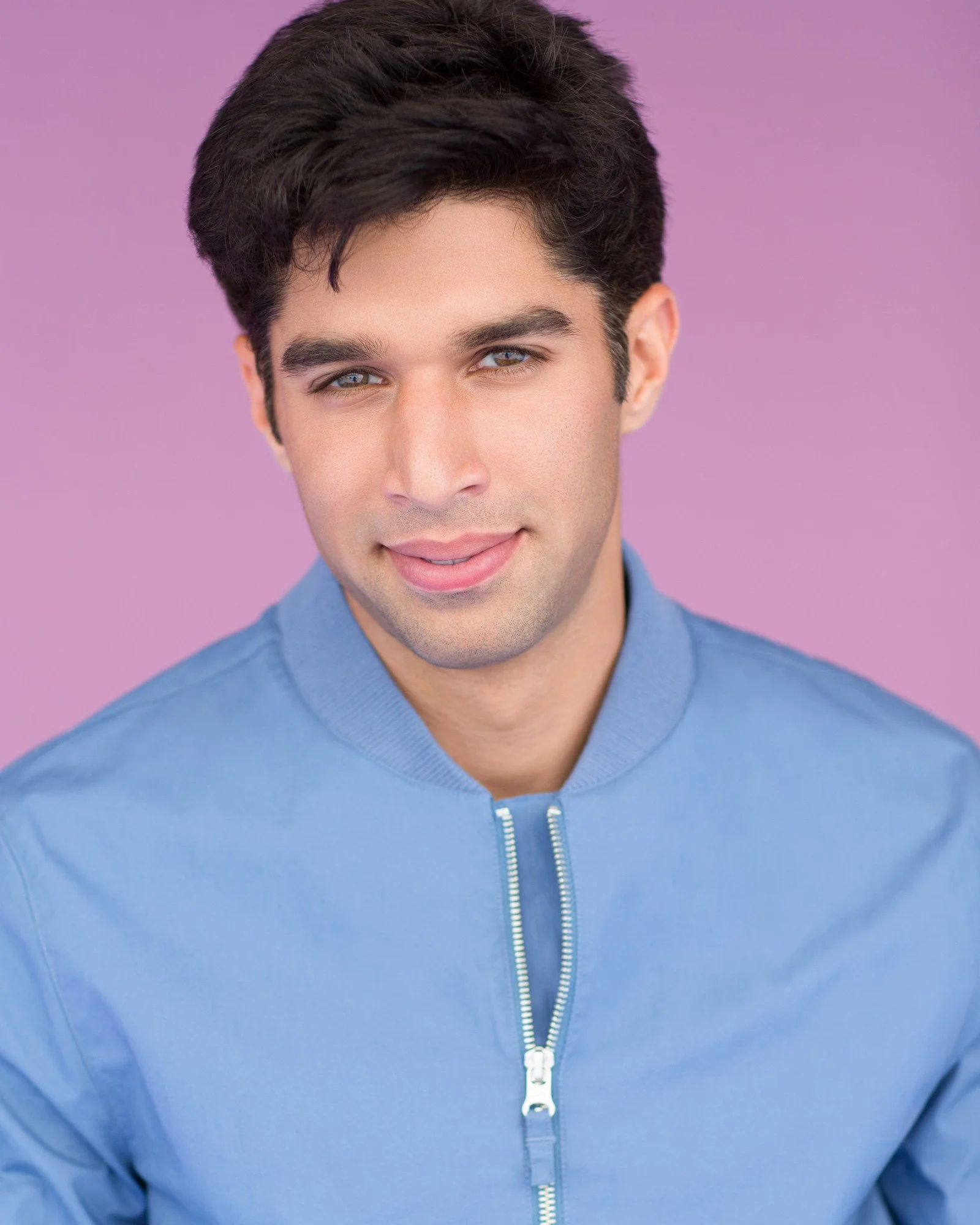 Close-up portrait of a young man with dark hair, blue eyes, and light skin, smiling slightly, wearing a light blue zip-up jacket against a purple background.