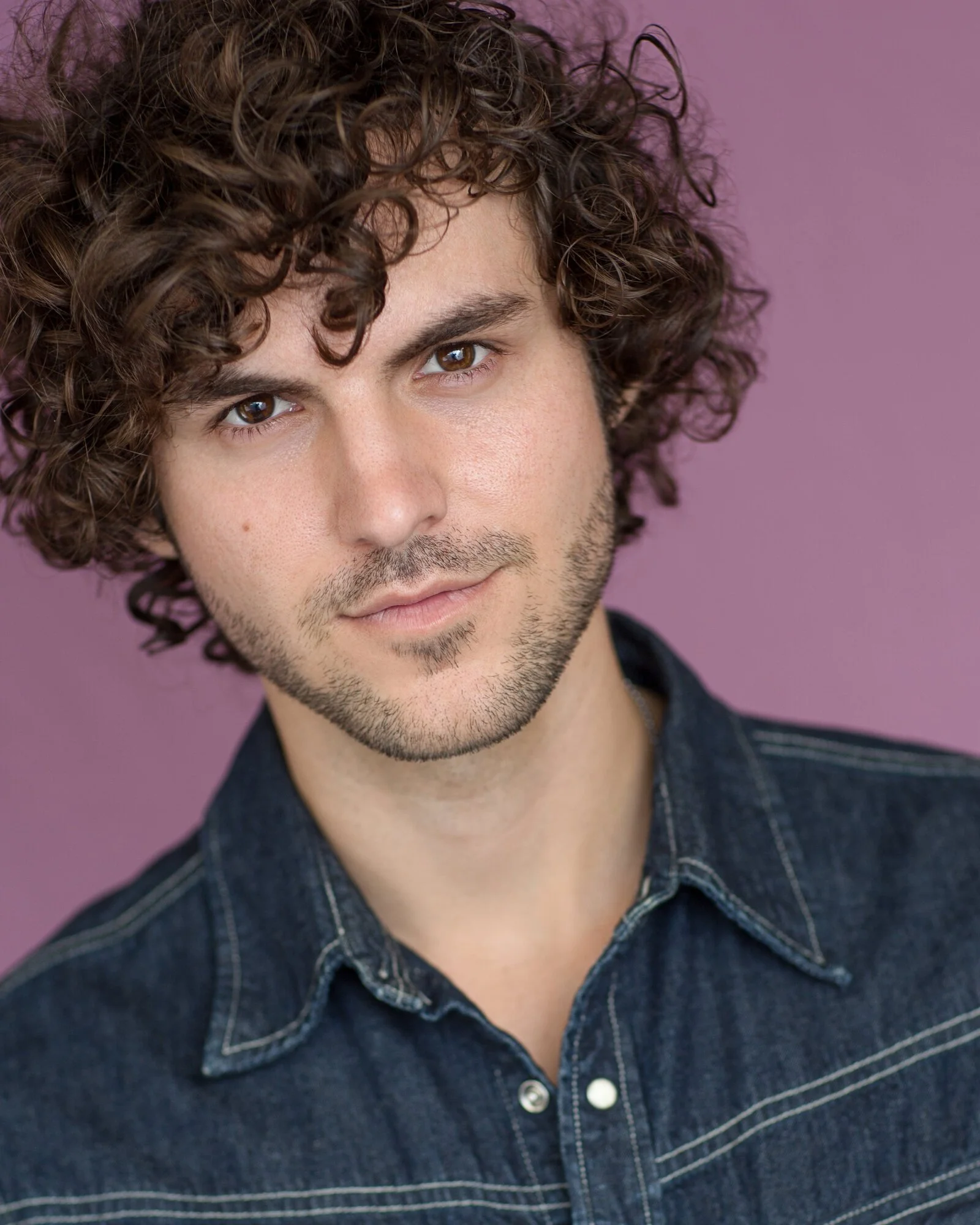 Close-up portrait of a young man with curly brown hair, brown eyes, light skin, and a slight beard, wearing a denim shirt, against a purple background.