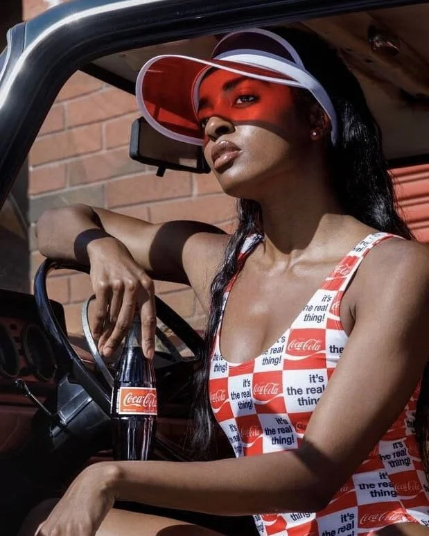 A woman wearing a red and white Coca-Cola swimsuit and a transparent visor hat, sitting in a vehicle with a brick wall in the background, holding a Coca-Cola glass bottle.