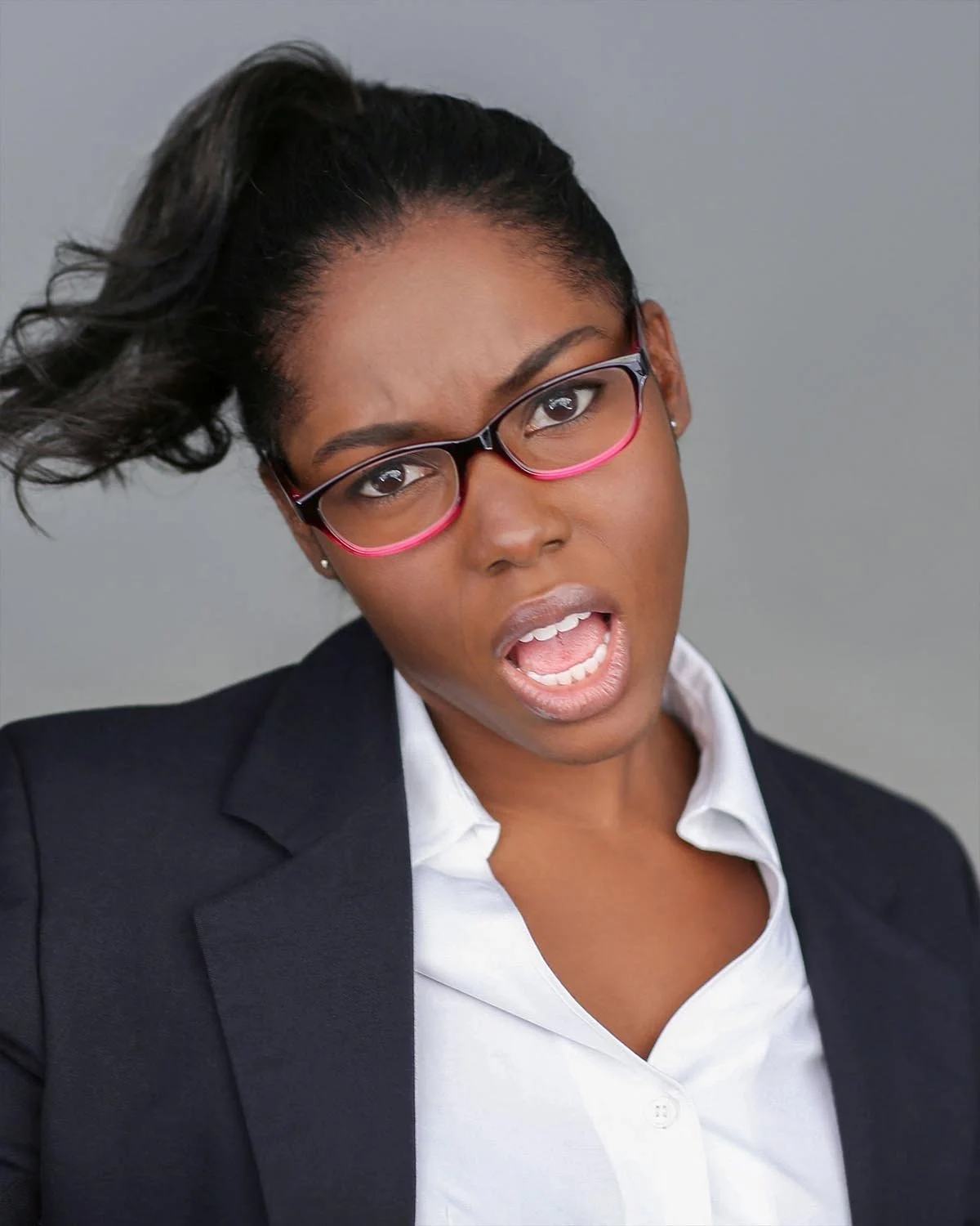 A young woman with glasses, wearing a black blazer and white shirt, making a surprised or confused facial expression against a plain gray background.