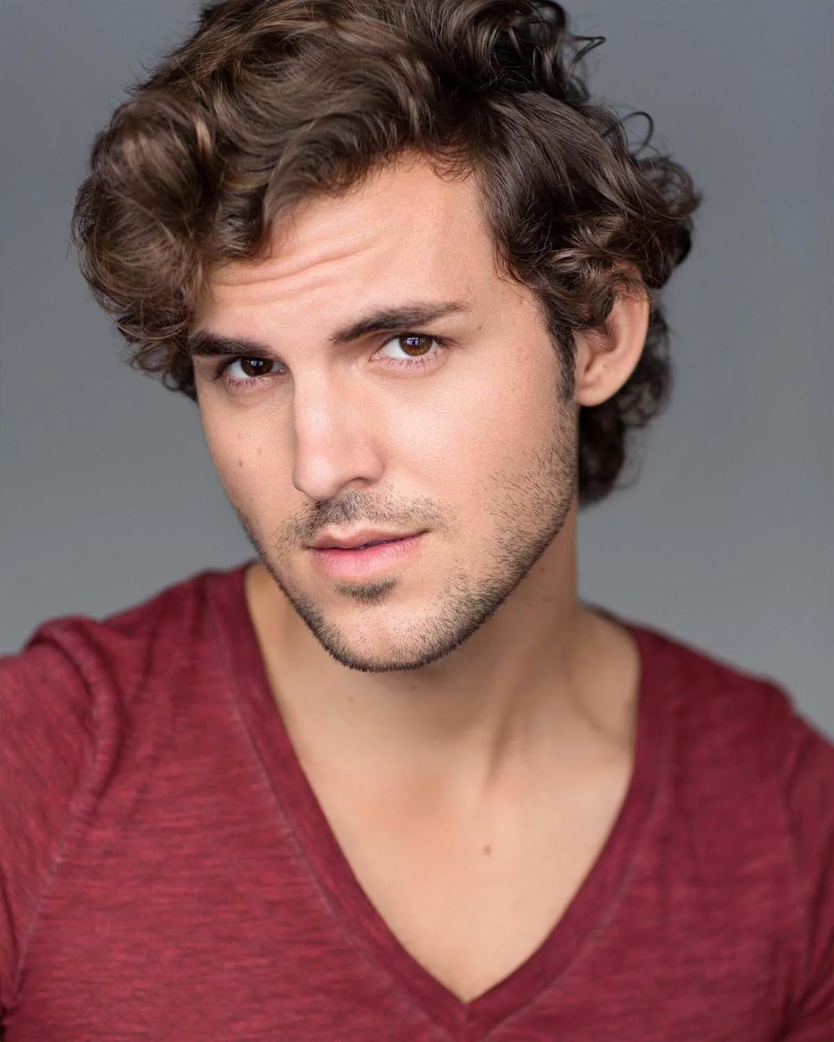 A young man with curly brown hair and light skin is wearing a red V-neck shirt and looking at the camera with a slightly serious expression.
