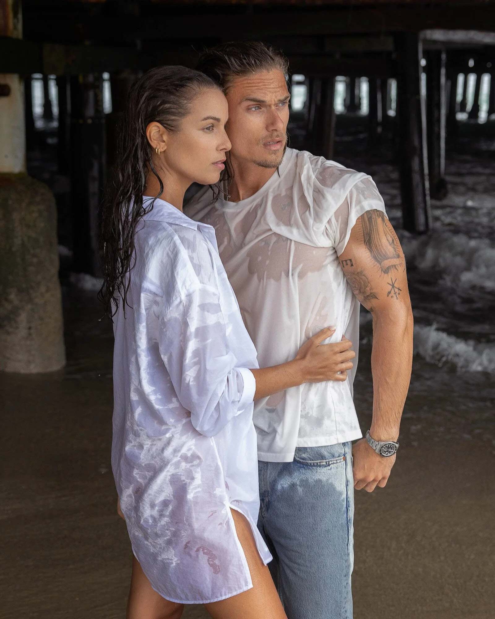 A man and woman are standing close together under a pier at the beach, both wearing wet white shirts, with the woman’s arm around the man’s waist.