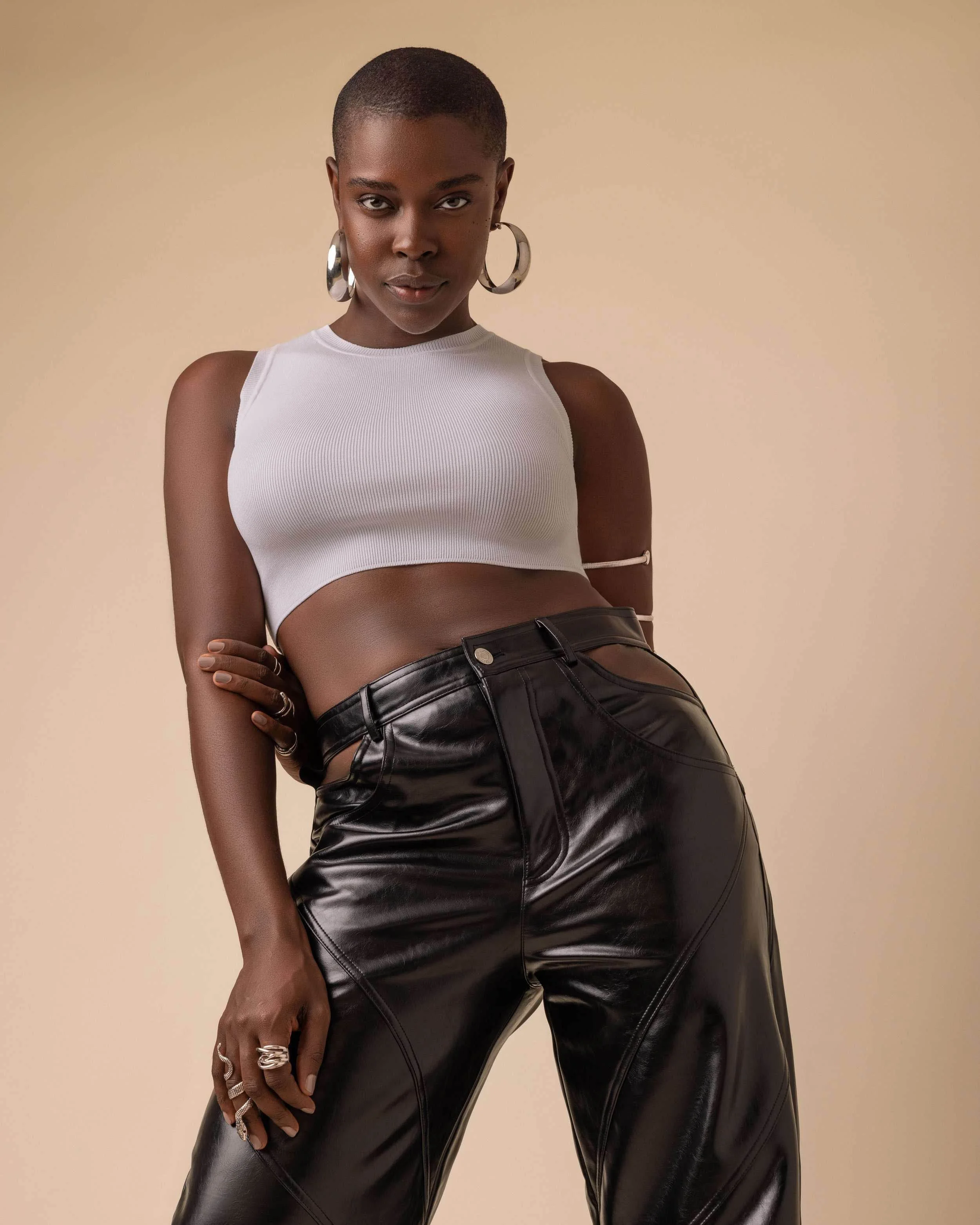 A confident black woman with a short haircut wearing a white sleeveless crop top and black leather pants, accessorized with large hoop earrings and rings, standing against a plain beige background.