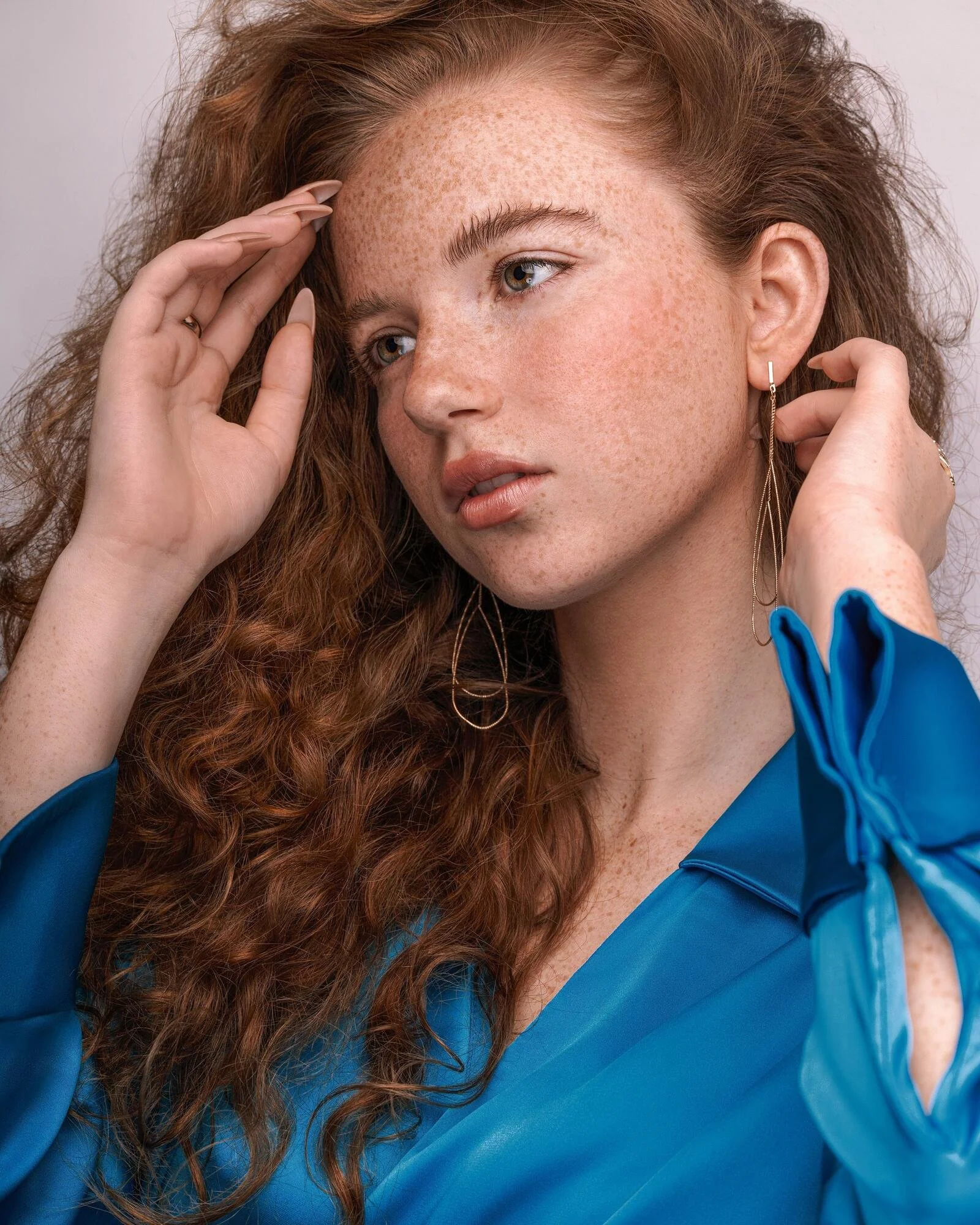 Close-up of a woman with long, curly red hair, fair skin with freckles, wearing a blue satin blouse and gold dangle earrings, posing with her hand near her temple.