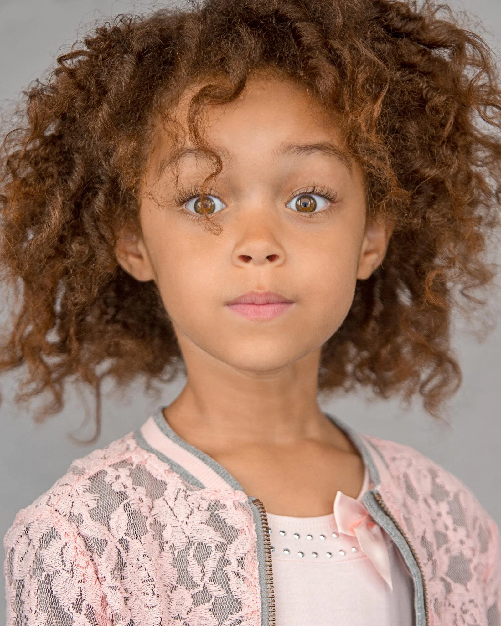 A young girl with curly brown hair and brown eyes looks directly at the camera. She is wearing a pink lace jacket over a light-colored top with small decorative elements and a small pink bow.