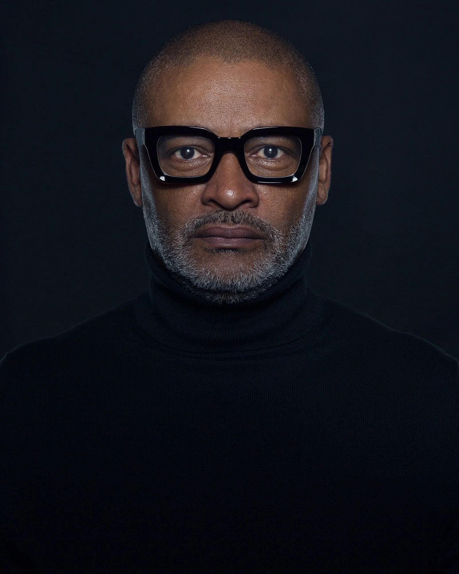 Portrait of a middle-aged man with gray beard and glasses, wearing a black turtleneck against a dark background.