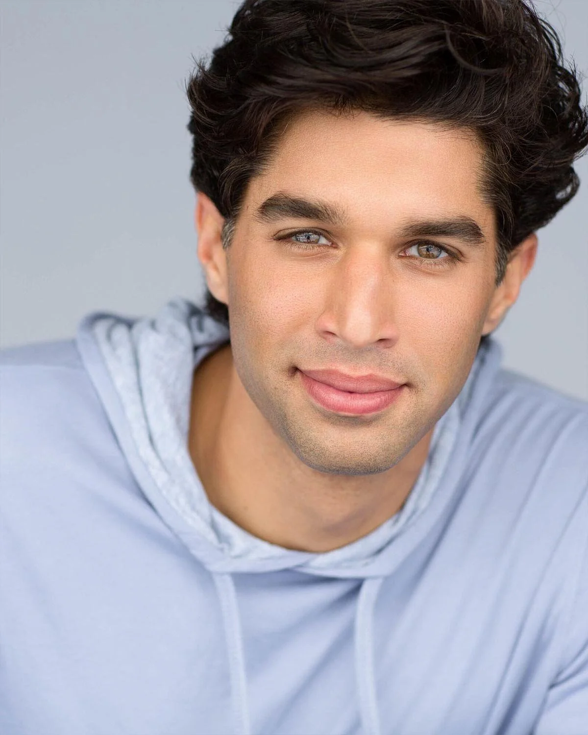 Close-up portrait of a young man with dark hair and blue eyes, smiling, wearing a light gray hoodie against a plain background.