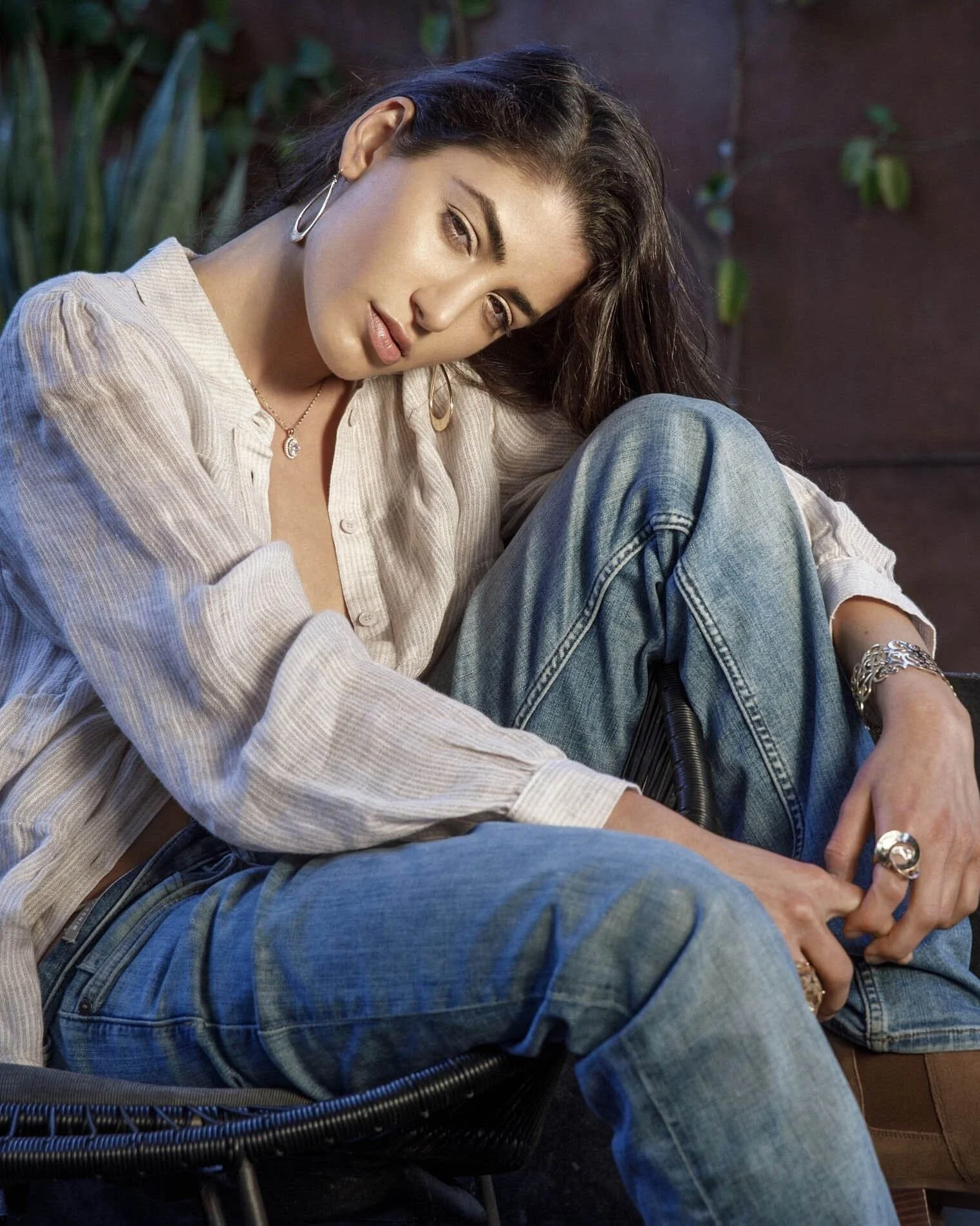 A woman with long dark hair, wearing earrings, a necklace, a linen shirt, and jeans, seated outdoors against a wooden wall with green plants in the background, looking at the camera.