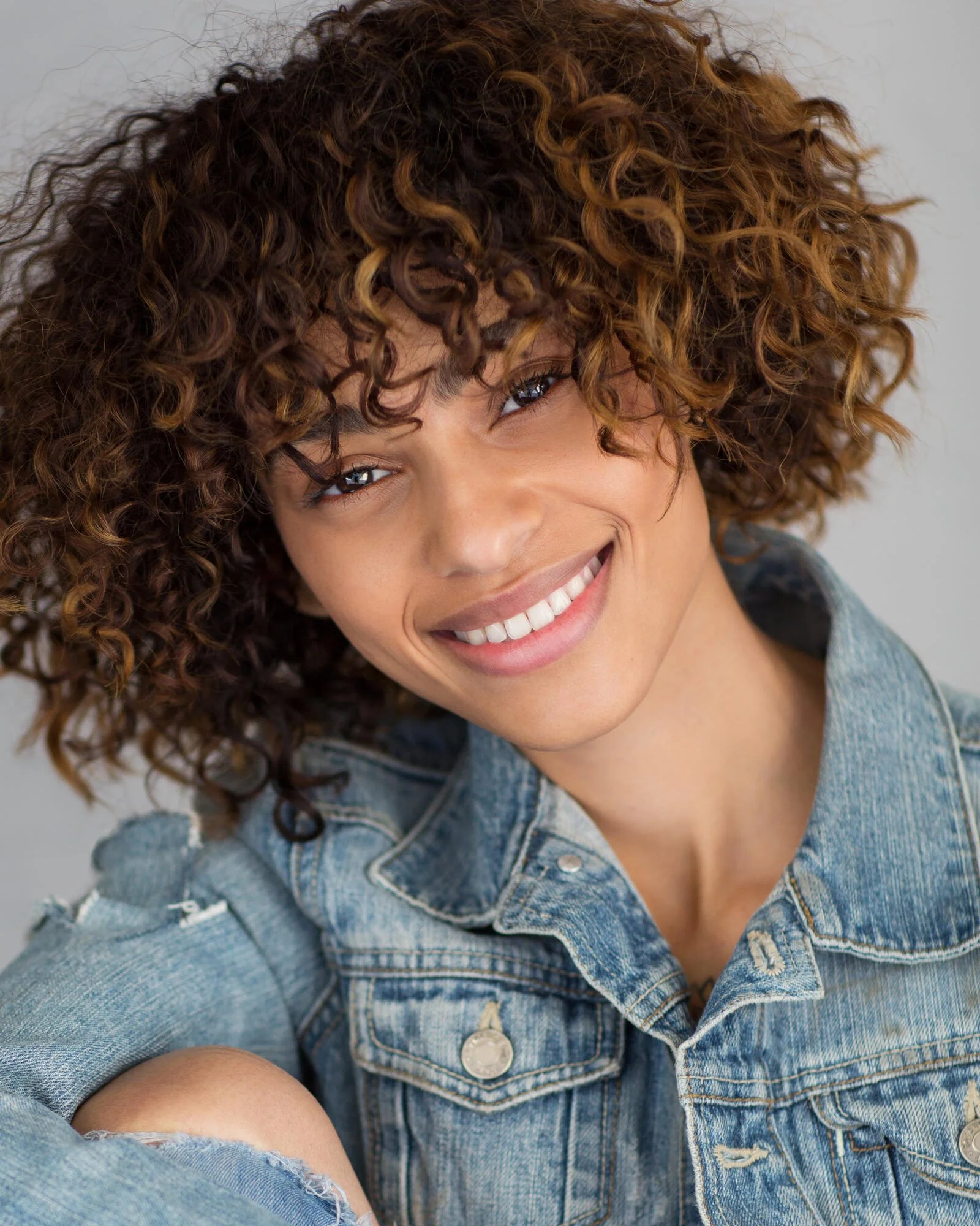 Close-up portrait of a woman with curly hair smiling, wearing a denim jacket.