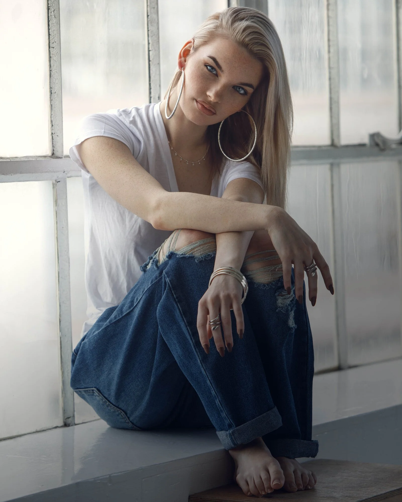A young woman with long blonde hair, wearing large hoop earrings, a white T-shirt, ripped jeans, multiple rings, bracelets, and a necklace, sitting on the floor near window panes with a natural light background.