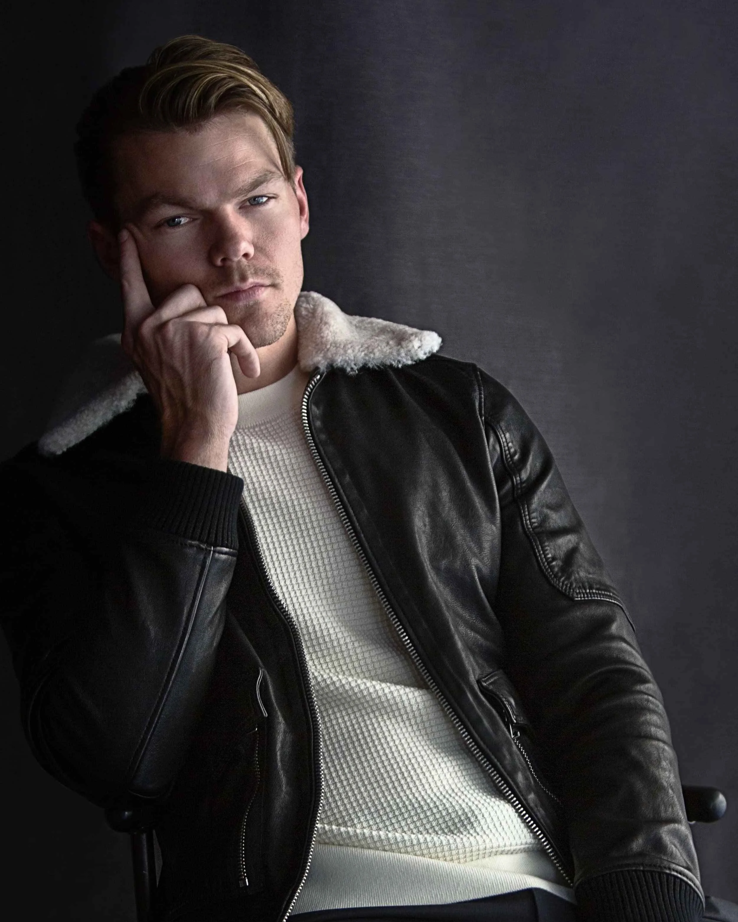 A young man with blonde hair and blue eyes, wearing a black leather jacket with a white fleece collar and a white shirt, posing against a dark background with his chin resting on his hand.