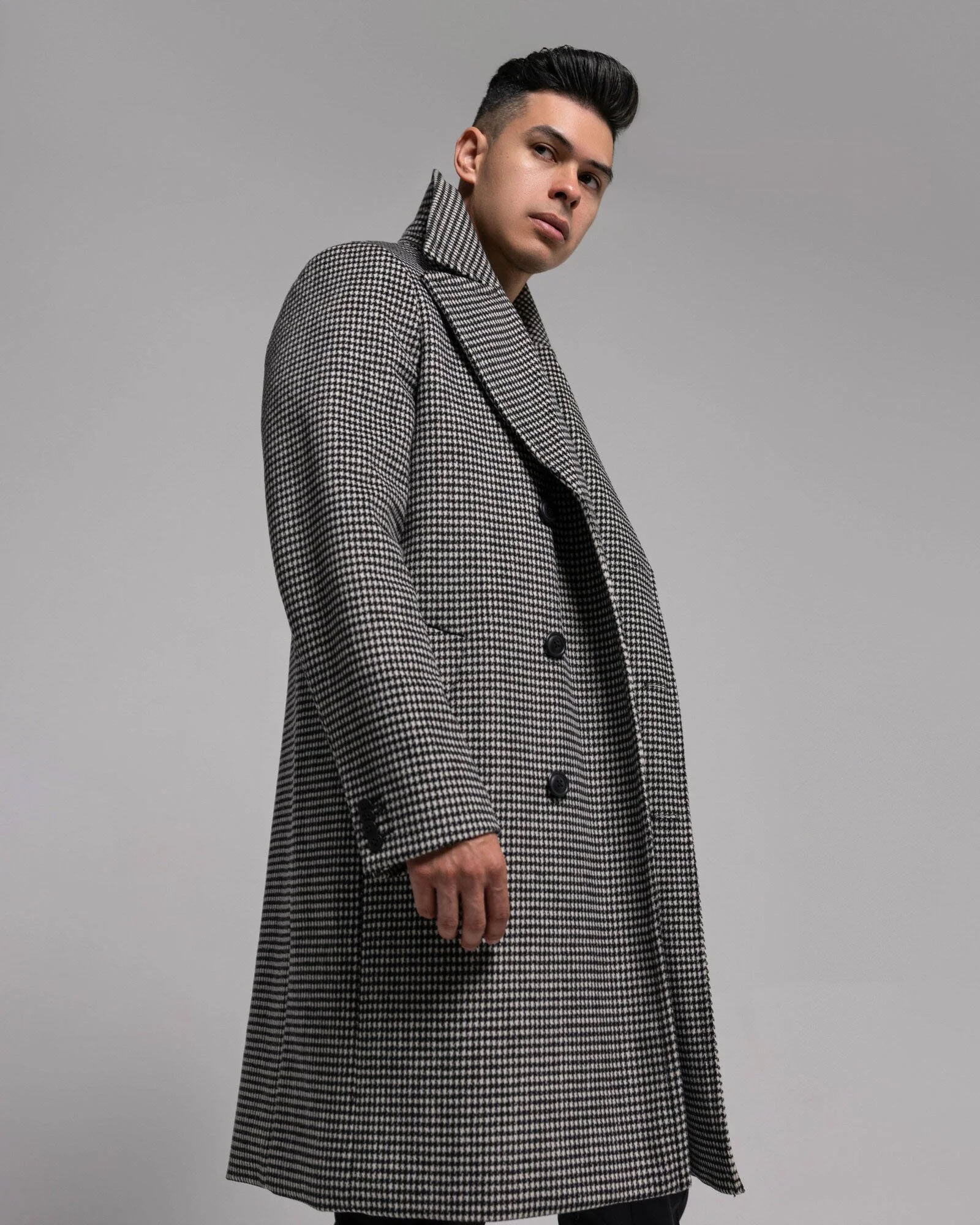 A young man with dark hair and a pensive expression, wearing a black-and-white houndstooth-patterned overcoat, standing against a plain gray background.