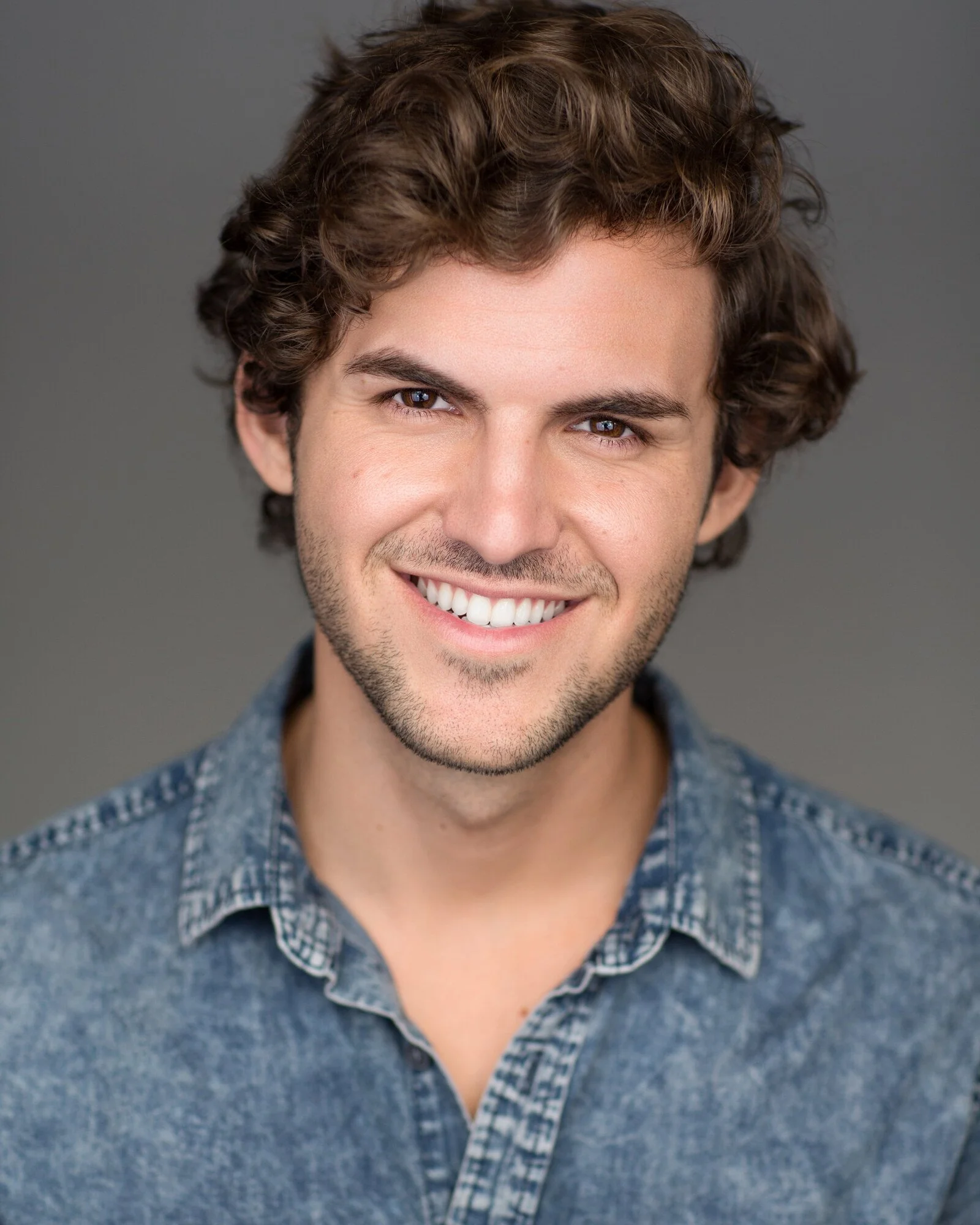 A smiling young man with curly brown hair, wearing a denim shirt, against a gray background.