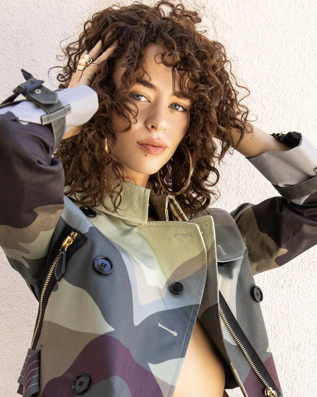 A woman with curly brown hair and hoop earrings posing against a plain wall, wearing a camouflage-patterned jacket.