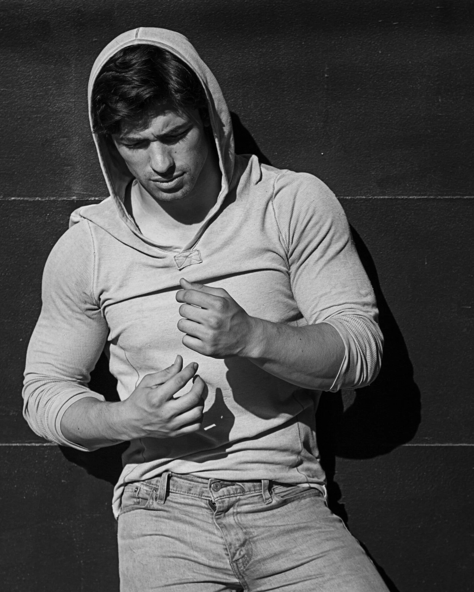 A young man leaning against a wall, wearing a hoodie and jeans, looking down with a serious expression.