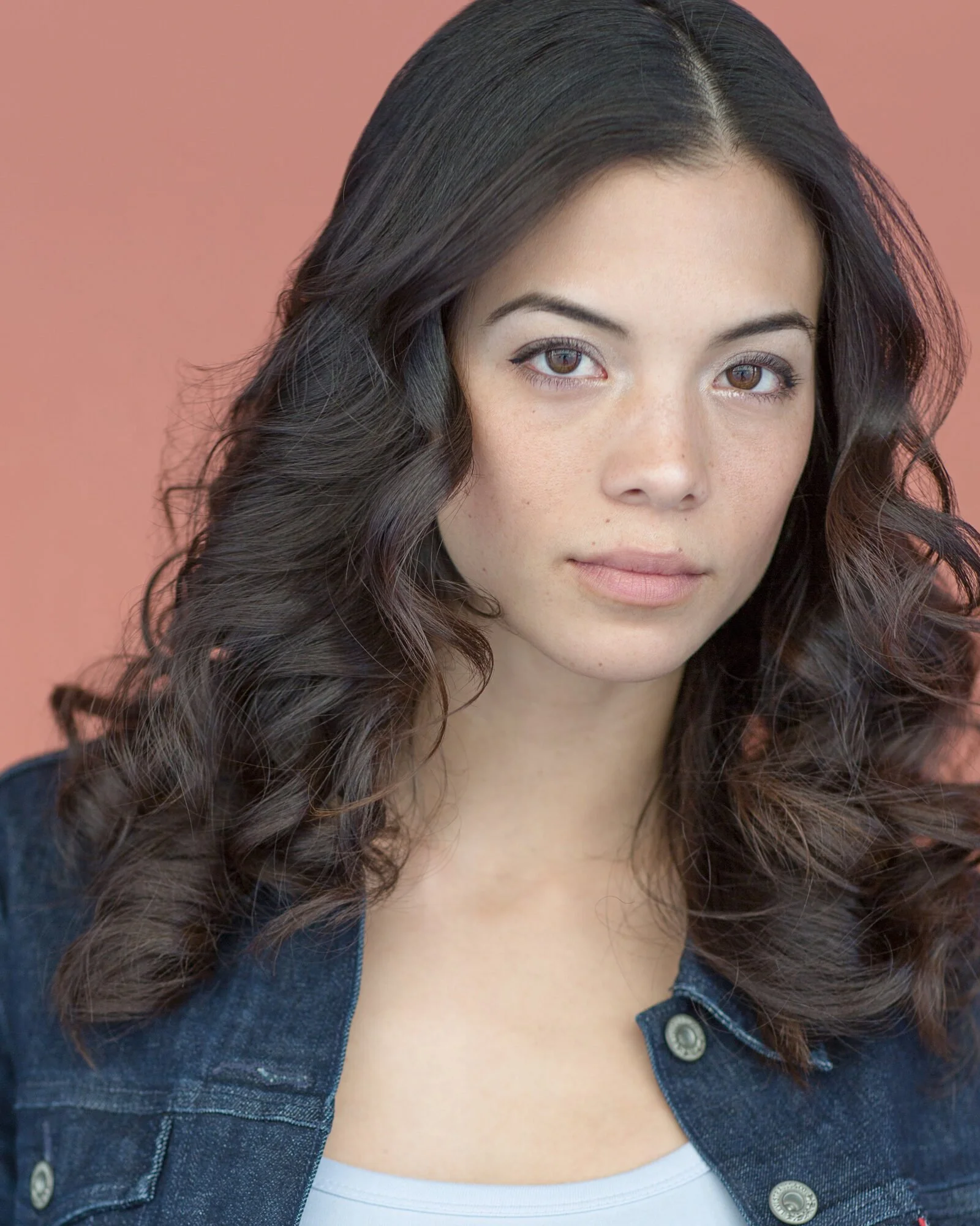 A young woman with long, dark, curly hair and light skin, looking directly at the camera, wearing a denim jacket and a white top, with a plain pink background.