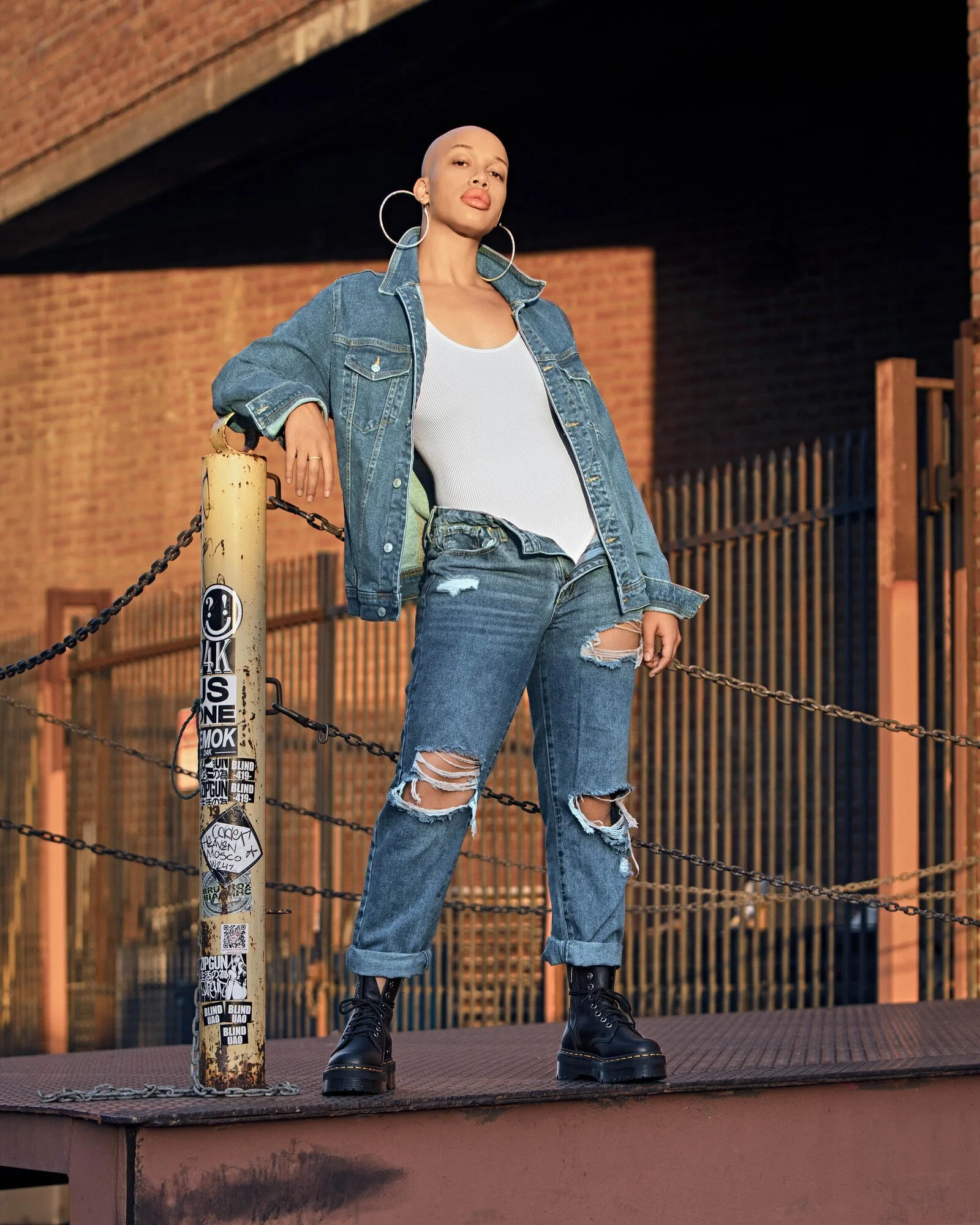 A woman with a shaved head wearing a denim jacket, white tank top, ripped jeans, black combat boots, standing on a skate park ramp, leaning on a chain barrier, with a brick building and fence in the background.