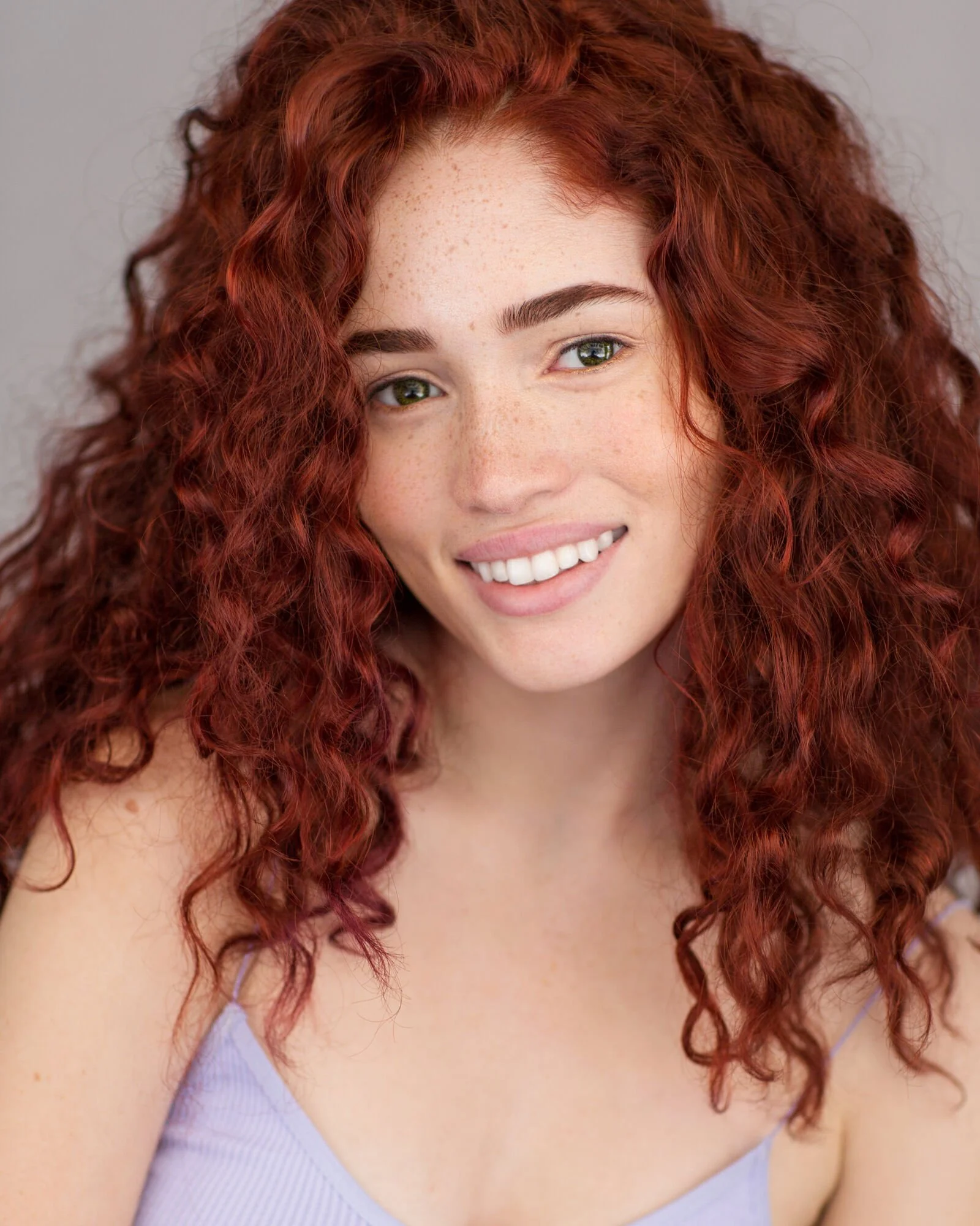 Close-up of a woman with curly red hair, green eyes, light freckles, and a bright smile