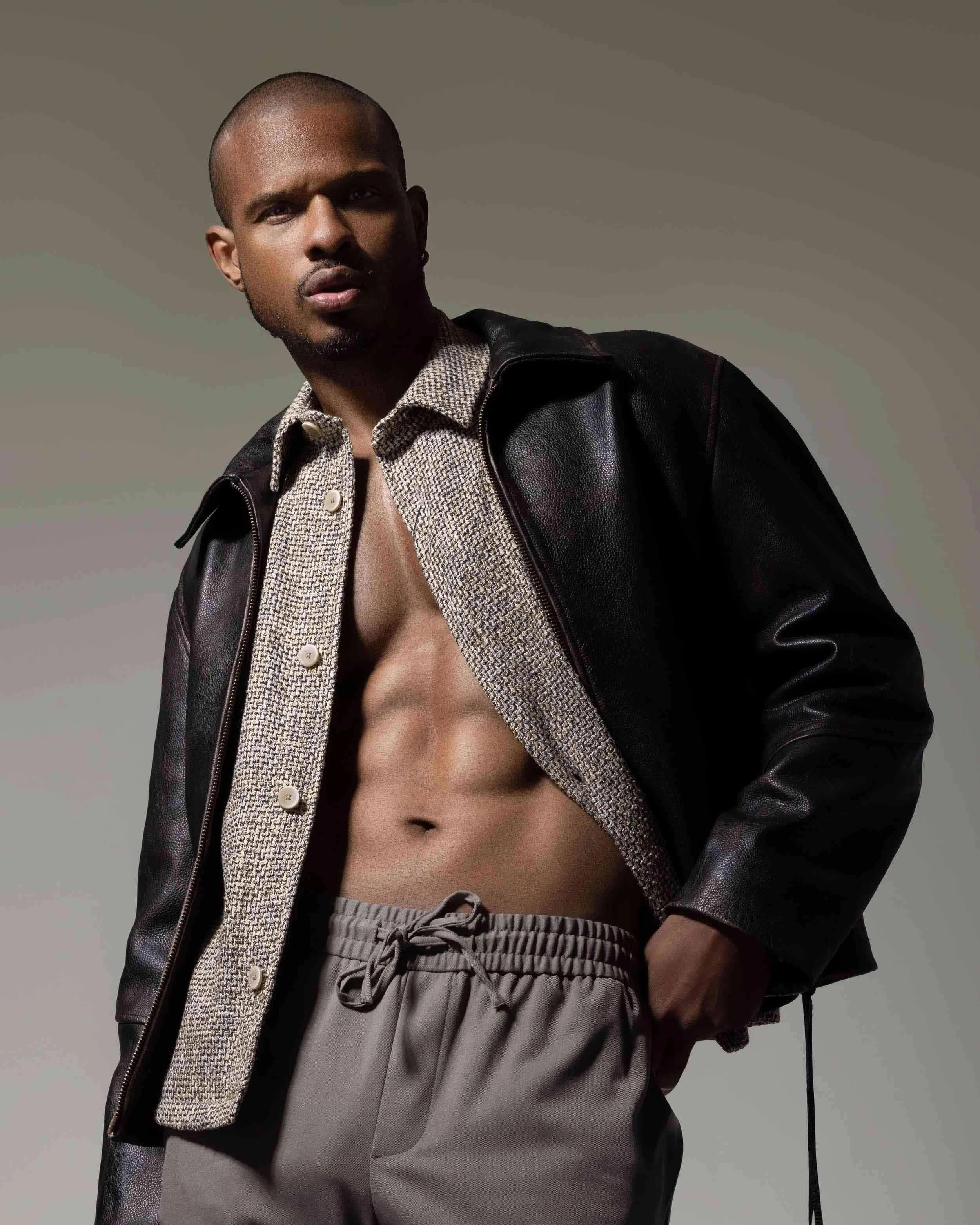Man with a shaved head and serious expression, wearing a black leather jacket, a partially unbuttoned patterned shirt revealing his toned abs, and gray sweatpants with a drawstring.