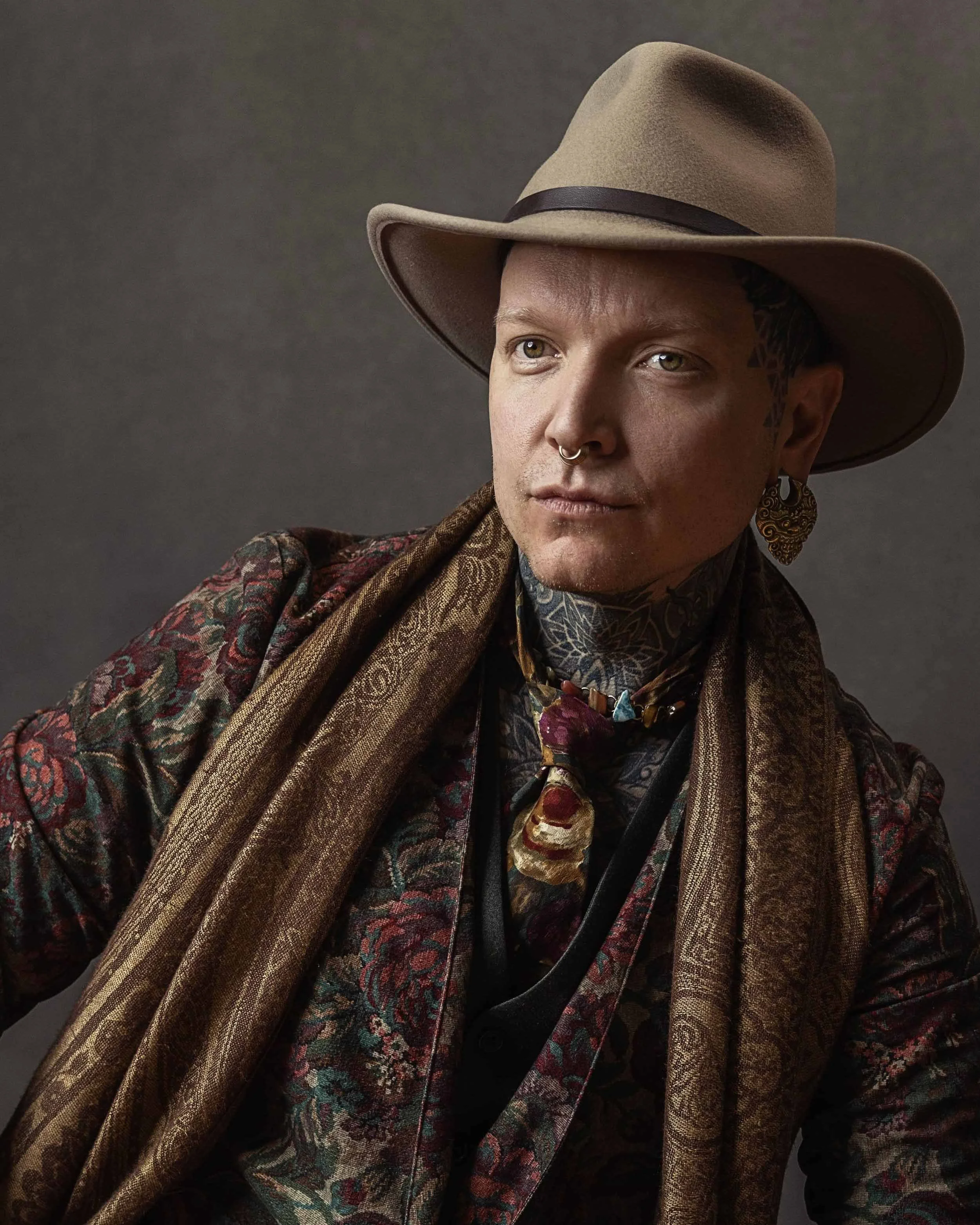 A man wearing a wide-brimmed tan hat, patterned blazer, and layered jewelry, including earrings and a scarf, against a plain background.