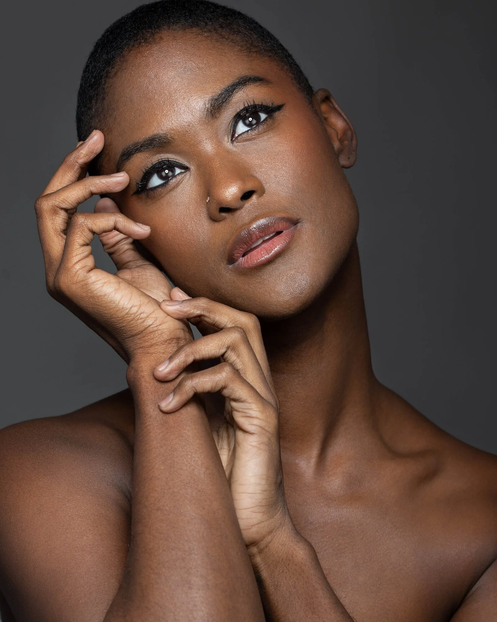 Close-up portrait of an African American woman with short hair, wearing winged eyeliner and neutral lipstick, touching her face with both hands against a dark gray background.