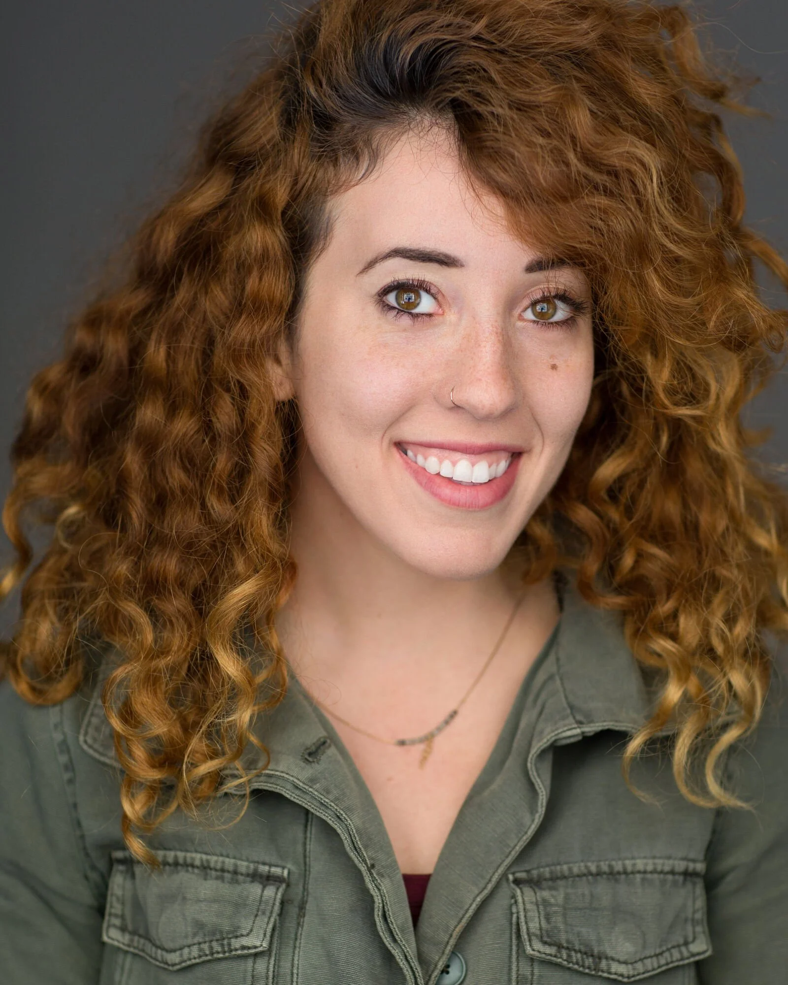 A young woman with curly red hair, light skin, and brown eyes, smiling and wearing a green jacket and necklace, against a gray background.