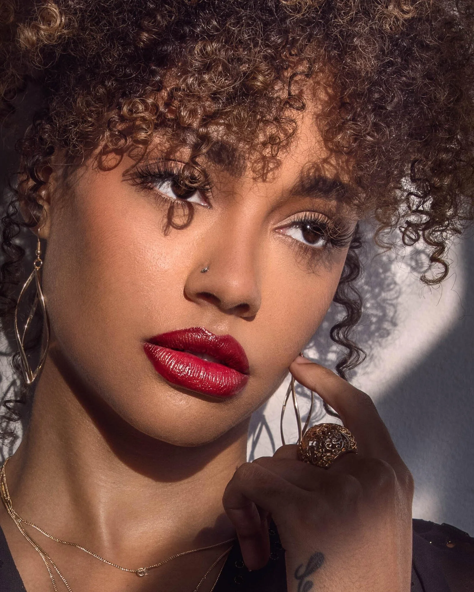 A woman with curly hair, red lipstick, gold earrings, and a nose piercing, holding her finger near her face and looking to the side.