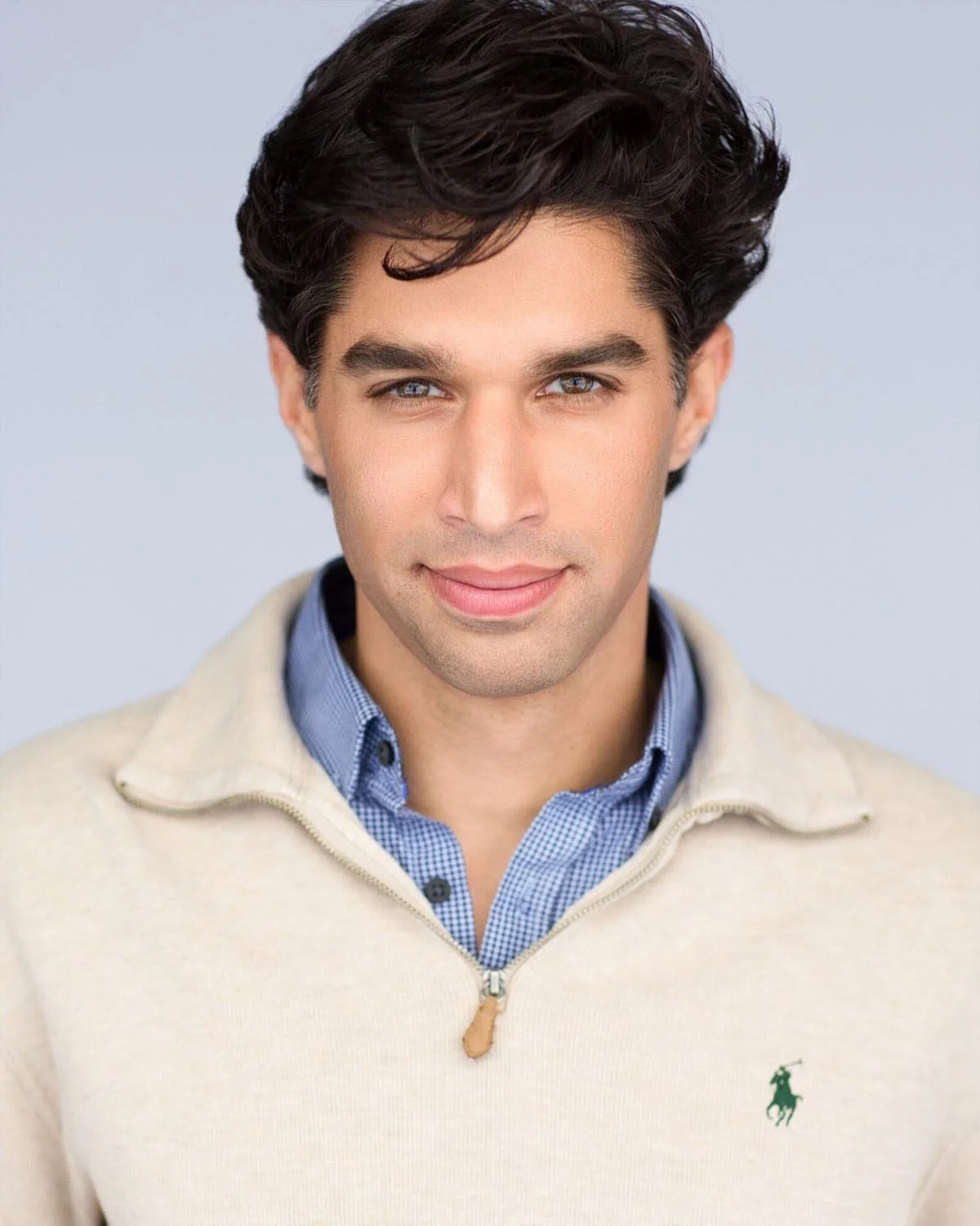 A young man with dark wavy hair and blue eyes smiling slightly, wearing a cream-colored quarter-zip sweater with a small green logo on the chest, over a blue checkered collared shirt, against a light gray background.