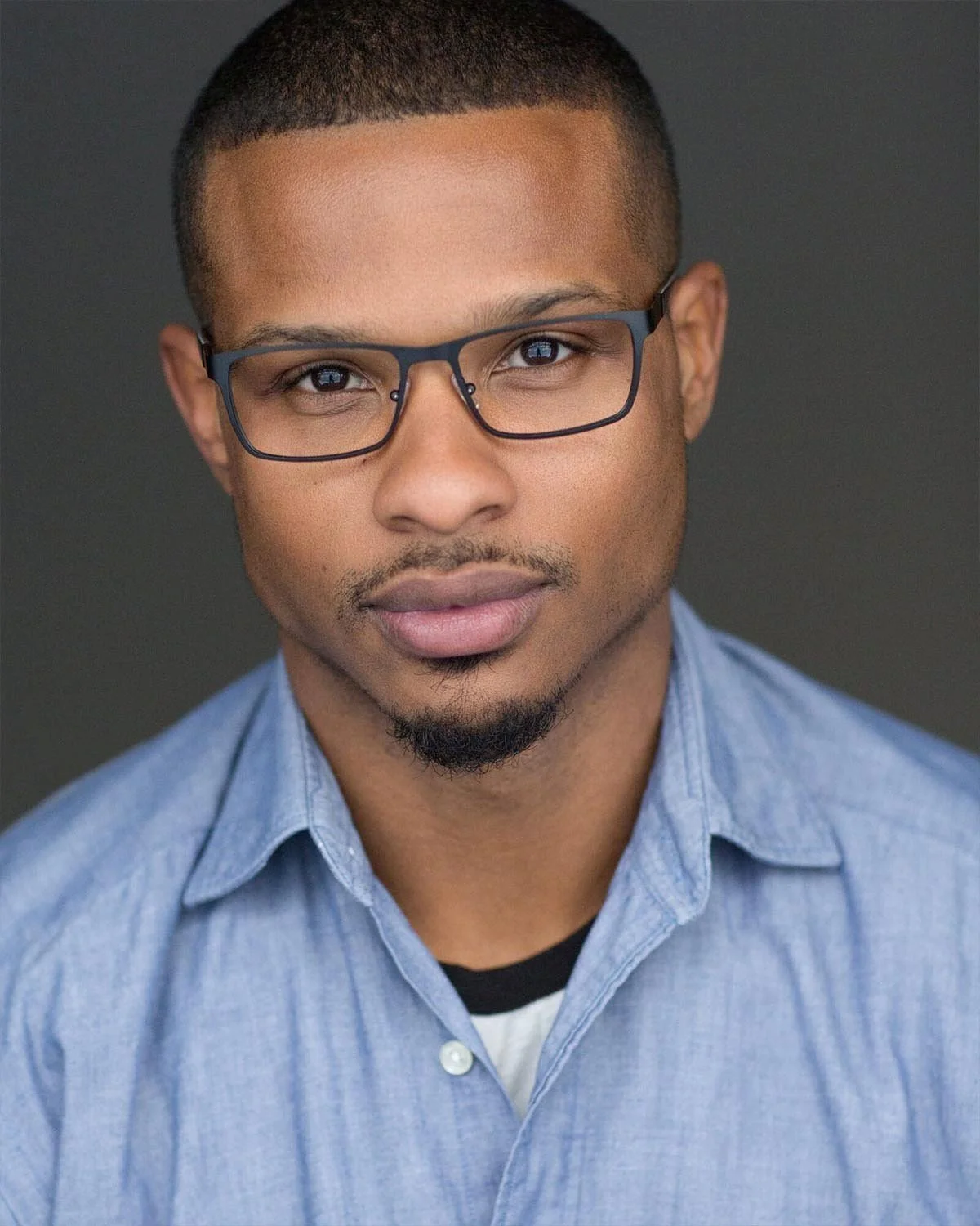 Close-up portrait of a young man wearing glasses and a blue denim shirt, looking directly at the camera against a dark background.