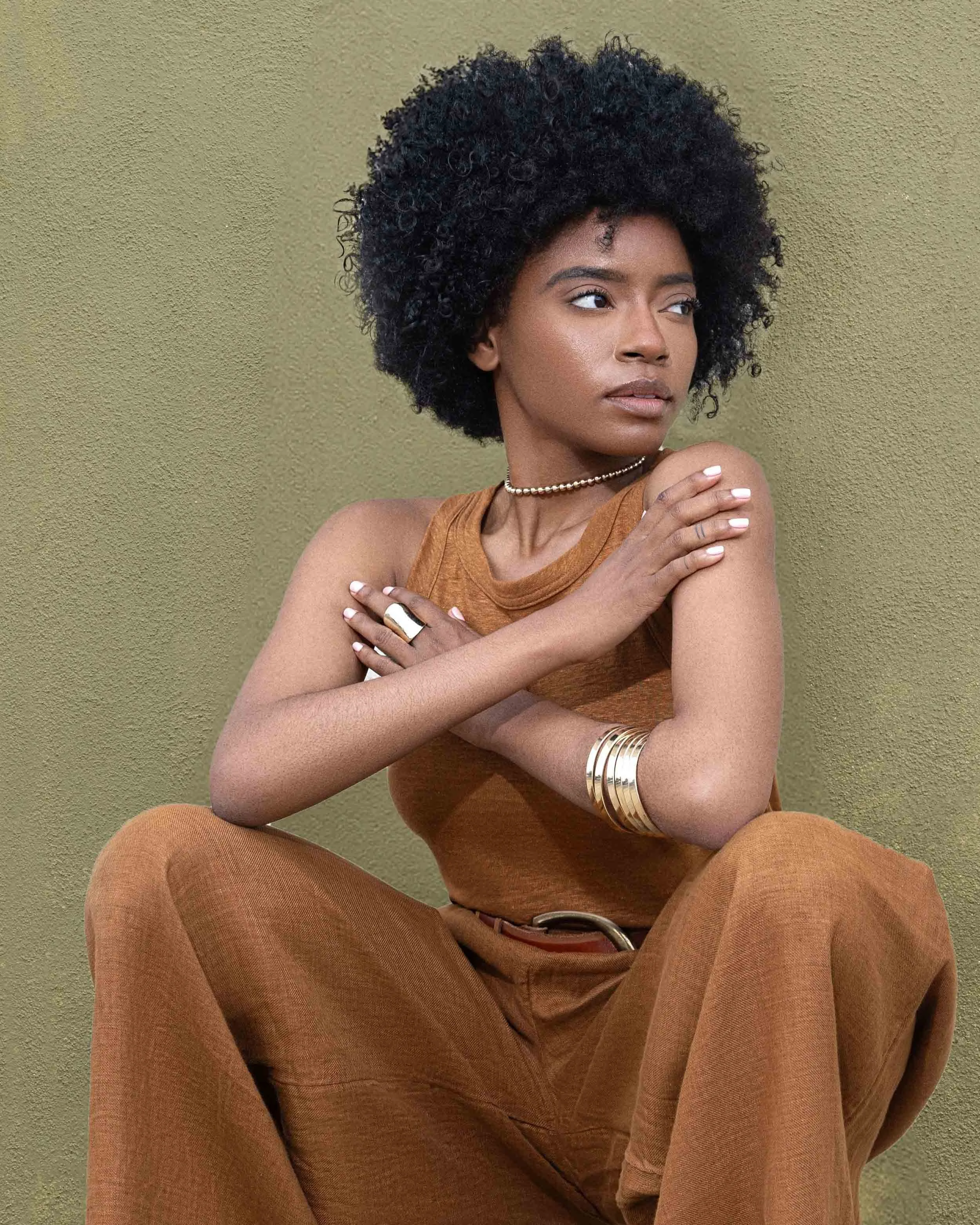 A young woman with curly black hair wearing a brown sleeveless top and matching brown pants, sitting in a relaxed pose against a plain textured beige background, with her arms crossed over her chest and looking to her right.