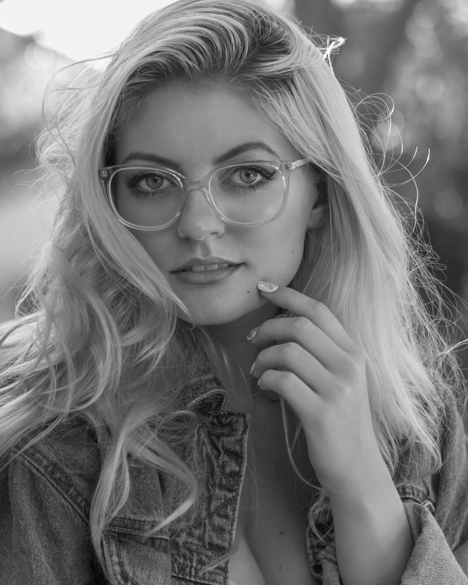 A portrait of a blonde woman with curly hair, wearing clear glasses, with her hand near her chin, wearing a denim jacket, outdoors, in black and white.