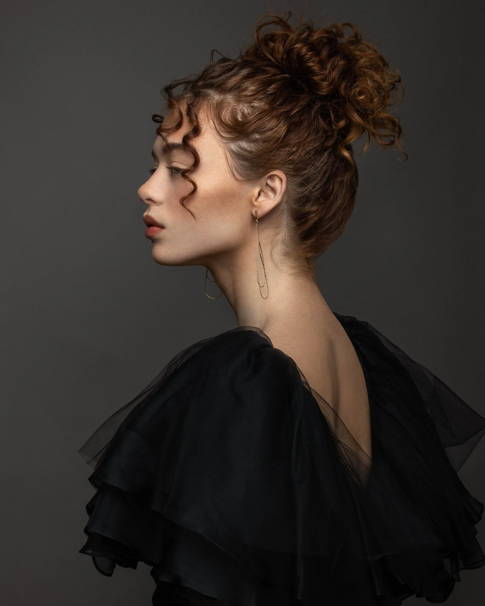 A woman with curly red hair styled in an updo, wearing long earrings and a black ruffled dress, positioned in profile against a dark background.